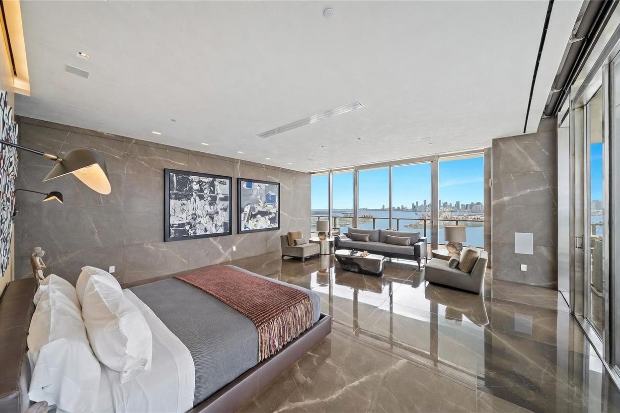 This expansive primary bedroom features a sophisticated, modern aesthetic with polished stone flooring and matching stone-clad walls. The room is anchored by a large bed with a dark frame and a textured throw, while a seating area with a sofa and two armchairs is positioned to take advantage of the panoramic water and city views through the large windows. The space is decorated with two large abstract art pieces, creating a luxurious and serene atmosphere.
