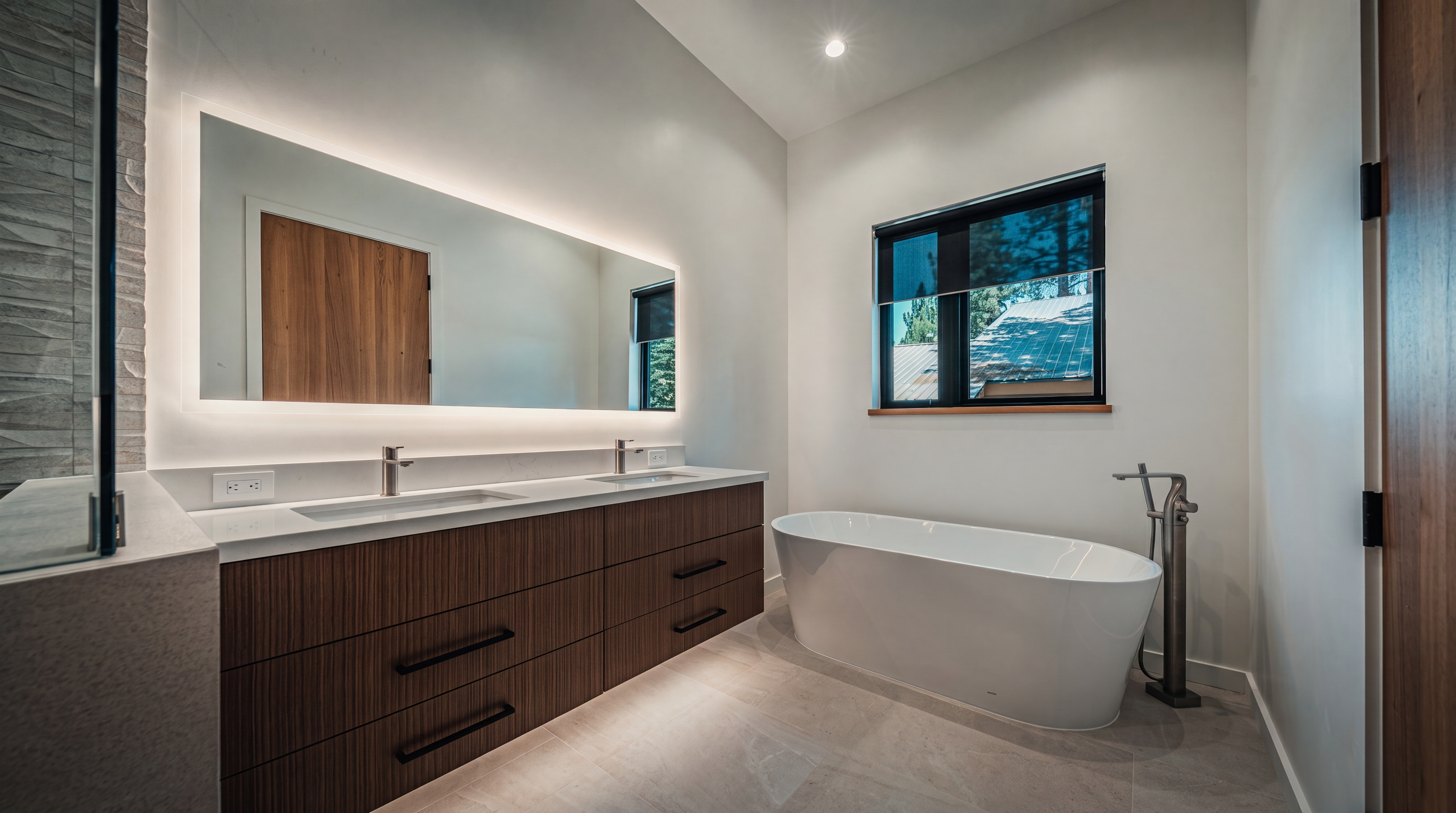 This modern primary bathroom features a sleek, floating dual-vanity with dark wood-toned cabinetry and a long, illuminated mirror. A freestanding white soaking tub sits prominently near a window, complemented by a tall, metallic floor-mounted faucet. The space is finished with light-toned tile flooring and clean, white walls, creating a bright and minimalist aesthetic.