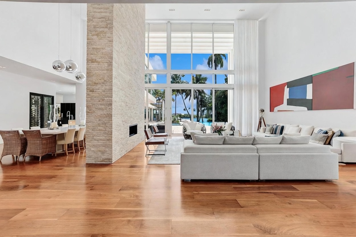 This expansive, open-concept living area features warm wood flooring and a prominent stone-clad chimney breast that anchors the space. Large, multi-pane windows provide an unobstructed view of the outdoor pool and tropical landscaping, bathing the room in natural light. The interior design is contemporary and airy, with a neutral color palette and a large sectional sofa positioned to enjoy both the indoor fireplace and the outdoor scenery.
