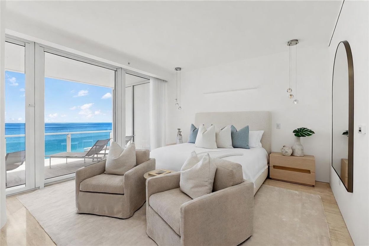 This bright and airy primary bedroom features a neutral color palette and a plush bed centered against a wall, flanked by light wood nightstands. Two comfortable armchairs are positioned in the foreground, creating a cozy seating area that faces the expansive floor-to-ceiling glass doors. These doors open onto a private balcony, offering a stunning, unobstructed view of the ocean and sky.