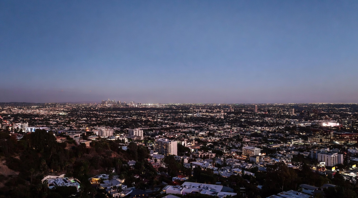 This sweeping aerial view captures a breathtaking panoramic vista of the Los Angeles cityscape at dusk. The perspective highlights the dense urban sprawl, illuminated by thousands of twinkling city lights, with the iconic downtown skyline visible in the distance. The foreground features lush hillside vegetation and residential rooftops, emphasizing the property's prime elevated location.