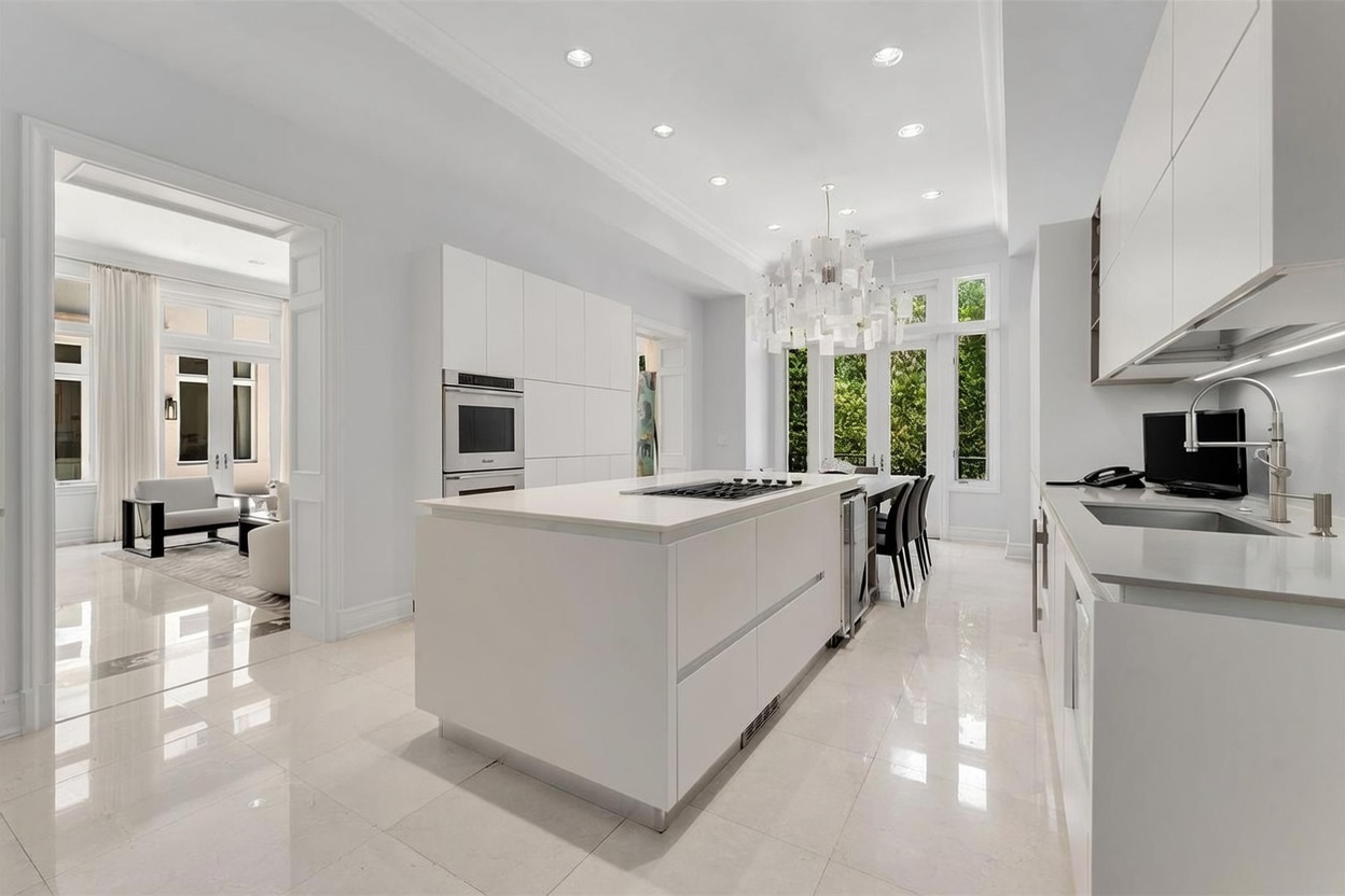 This bright, modern kitchen features a minimalist aesthetic with all-white cabinetry, a large central island with a built-in cooktop, and polished light-colored stone flooring. A striking, multi-tiered geometric chandelier hangs above the island, while large windows and glass doors provide ample natural light and a view into an adjacent living area. The space is designed with clean lines and high-end finishes, creating an airy and sophisticated atmosphere.