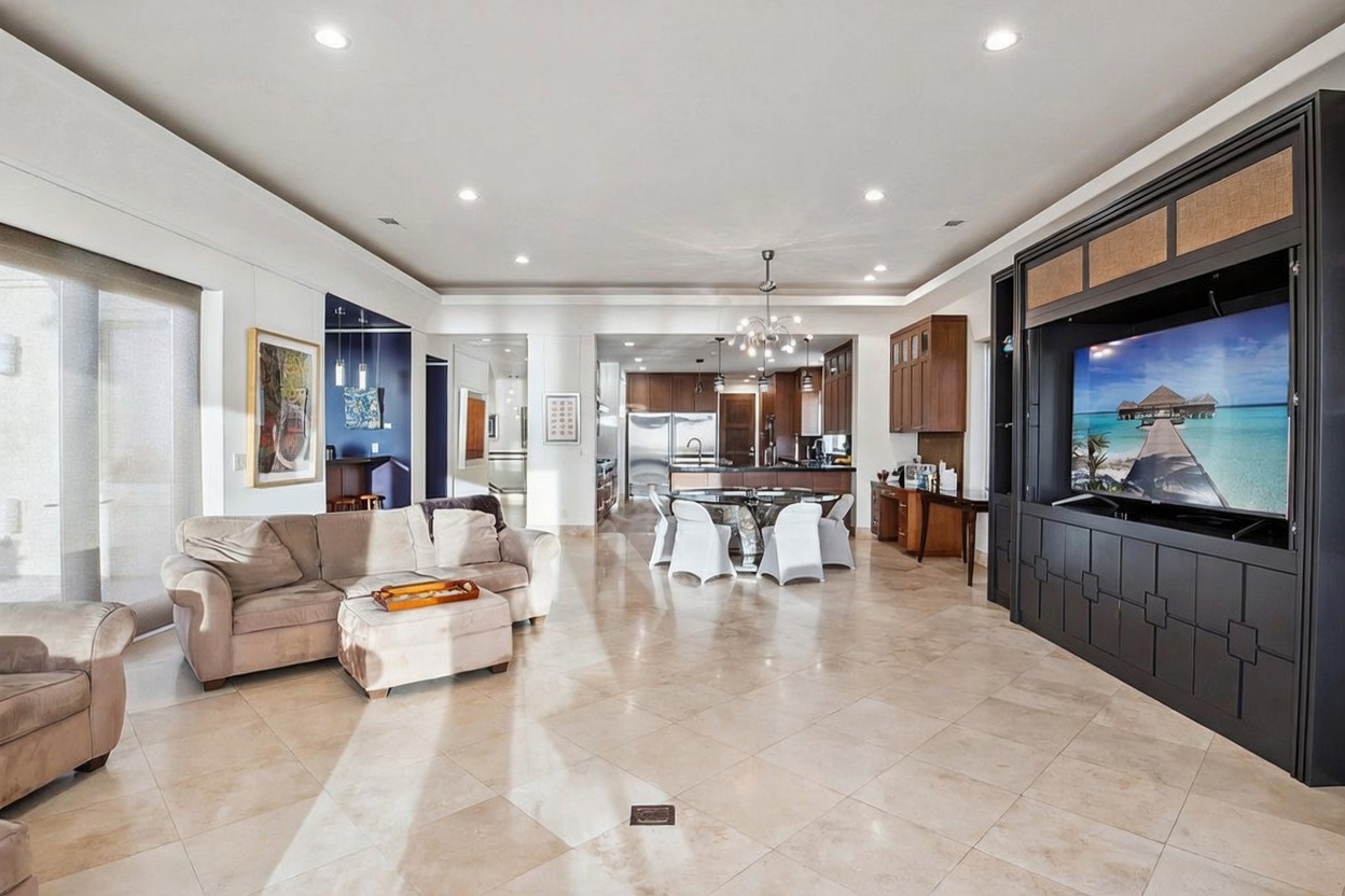 This spacious open-concept living area features light-toned tile flooring and a large, dark-toned built-in media center housing a television. A comfortable beige sofa and matching ottoman are positioned in the foreground, while a dining area with a glass-topped table and white chairs sits in the background near the kitchen. The perspective is a wide-angle shot from the living room looking toward the kitchen, showcasing the expansive, airy layout of the home.