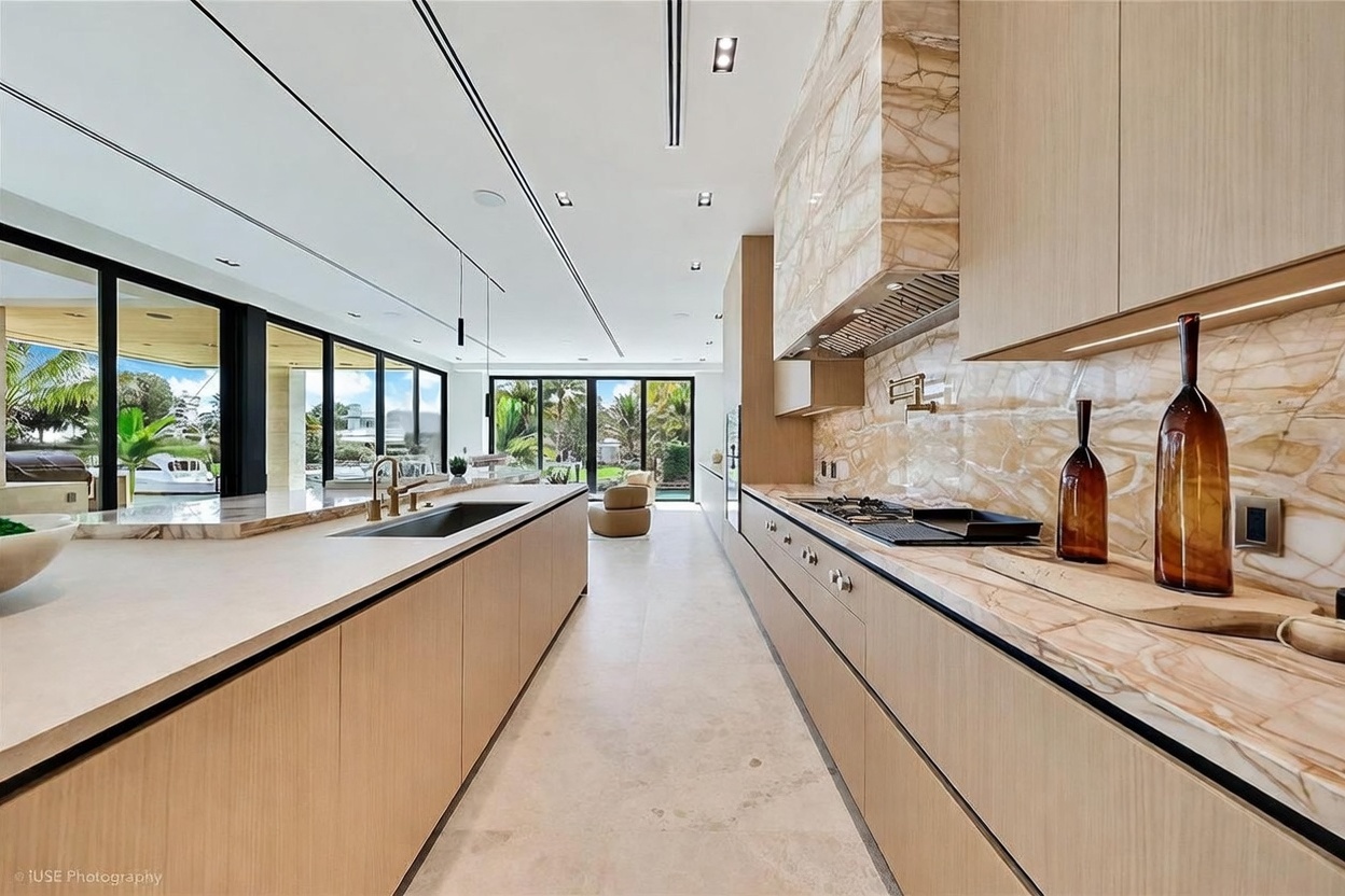 This sleek, modern kitchen features a long central island with a stone countertop and a large integrated sink, paired with light wood cabinetry. A striking stone backsplash and matching range hood create a cohesive, high-end aesthetic, while large glass doors at the end of the room provide a seamless transition to the outdoor space. The perspective is a wide-angle shot looking down the length of the kitchen, emphasizing the clean lines and open-concept layout.