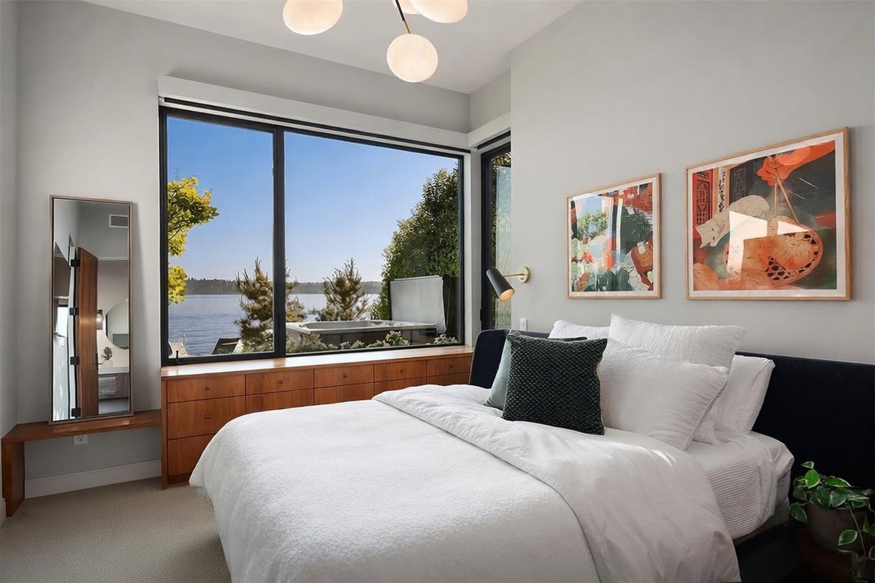 This serene bedroom features a large window offering a scenic view of the water and a private outdoor spa. The room is furnished with a bed dressed in white linens, a dark upholstered headboard, and a long wooden dresser beneath the window. Two framed art prints hang on the light-colored wall, and a modern multi-globe light fixture hangs from the ceiling, creating a bright and tranquil atmosphere.