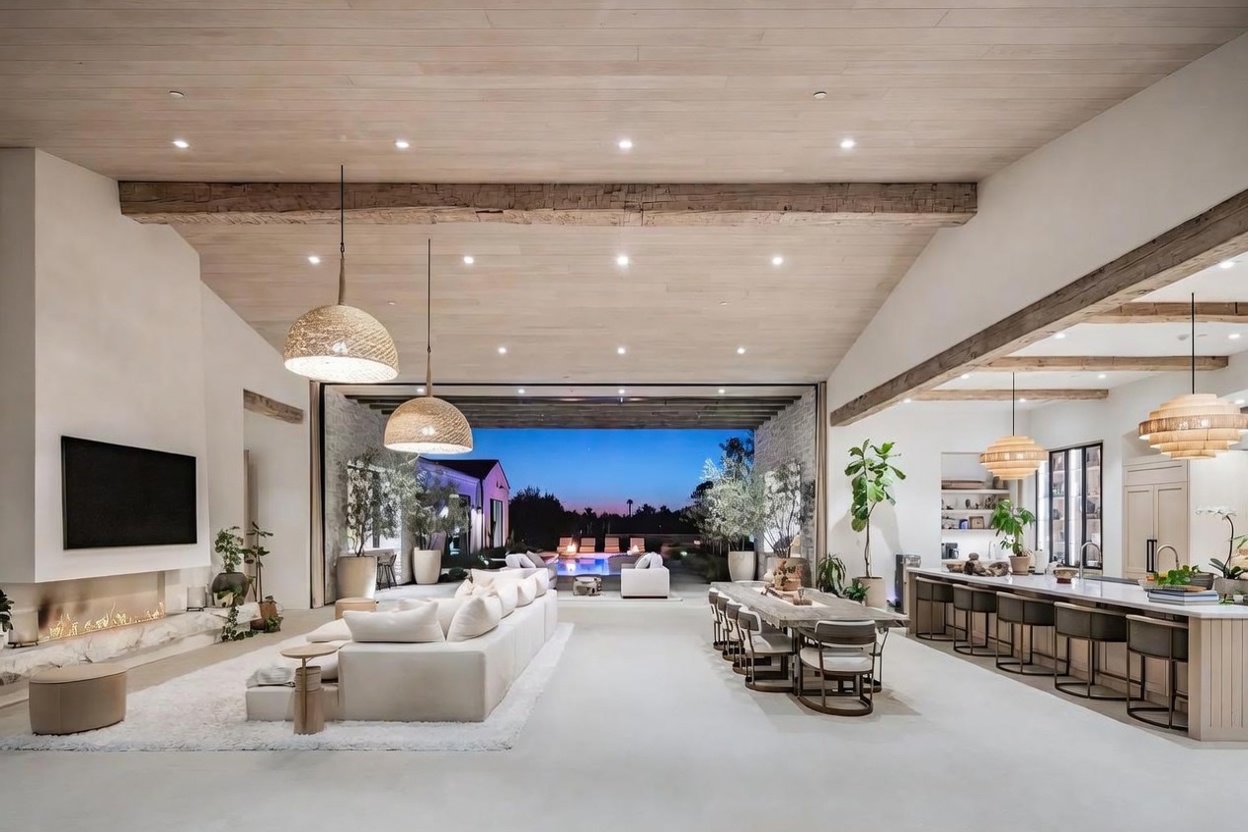 This expansive open-concept living area features a seamless transition to an outdoor pool deck through a wide, unobstructed opening. The space is defined by a light wood-paneled ceiling with exposed timber beams, a large white sectional sofa, and a long dining table, all illuminated by textured woven pendant lights. A stone-clad chimney breast anchors the living area, creating a sophisticated, airy atmosphere that emphasizes indoor-outdoor living.