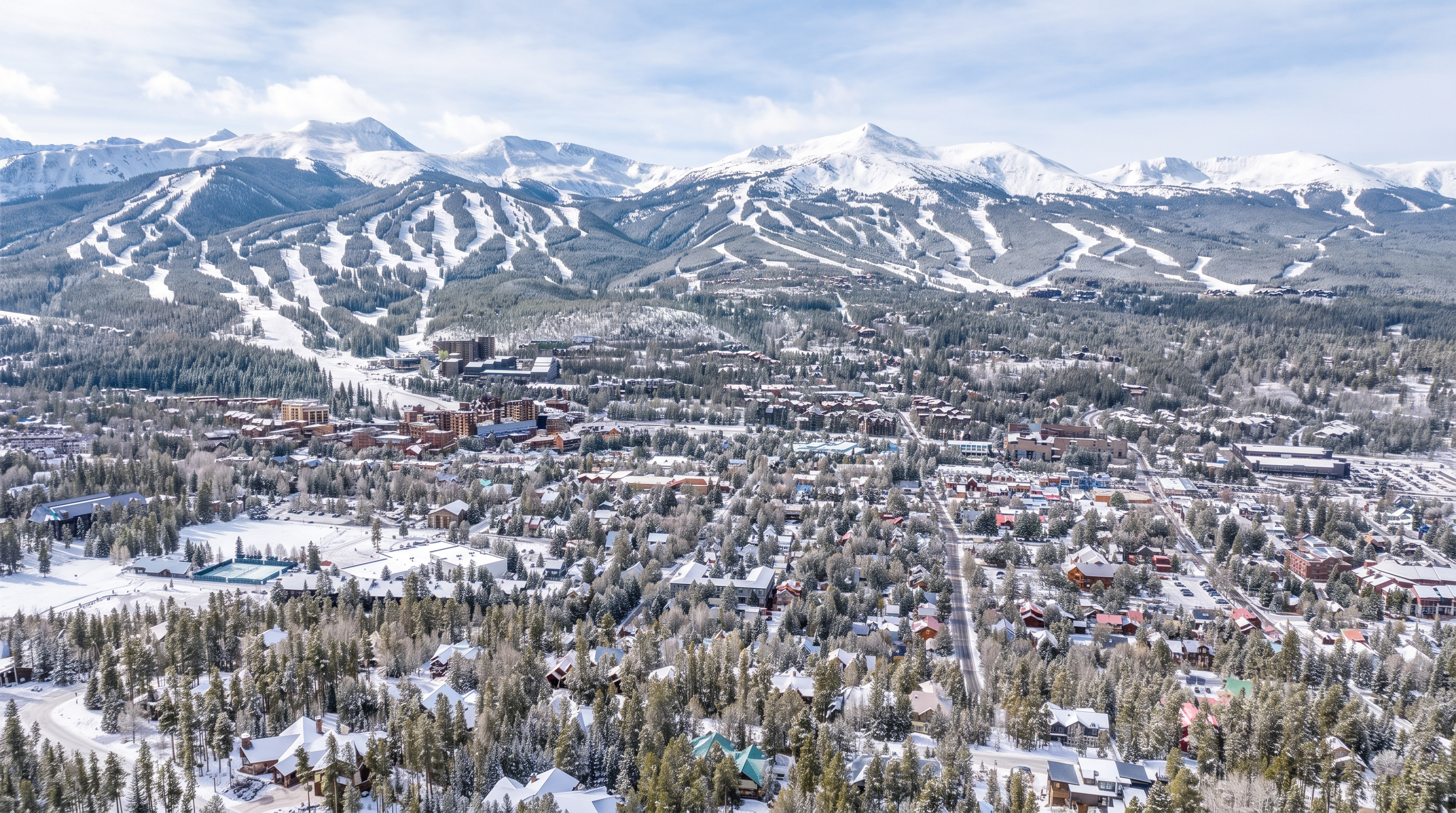 This high-angle aerial view captures a sprawling mountain town nestled at the base of a prominent ski resort. The landscape is characterized by dense evergreen forests, winding residential streets, and a mix of mountain-style homes, with the dramatic, rugged peaks of the ski slopes dominating the background under a bright, cloudy sky. The perspective provides a comprehensive overview of the community's layout and its immediate proximity to outdoor recreational terrain.