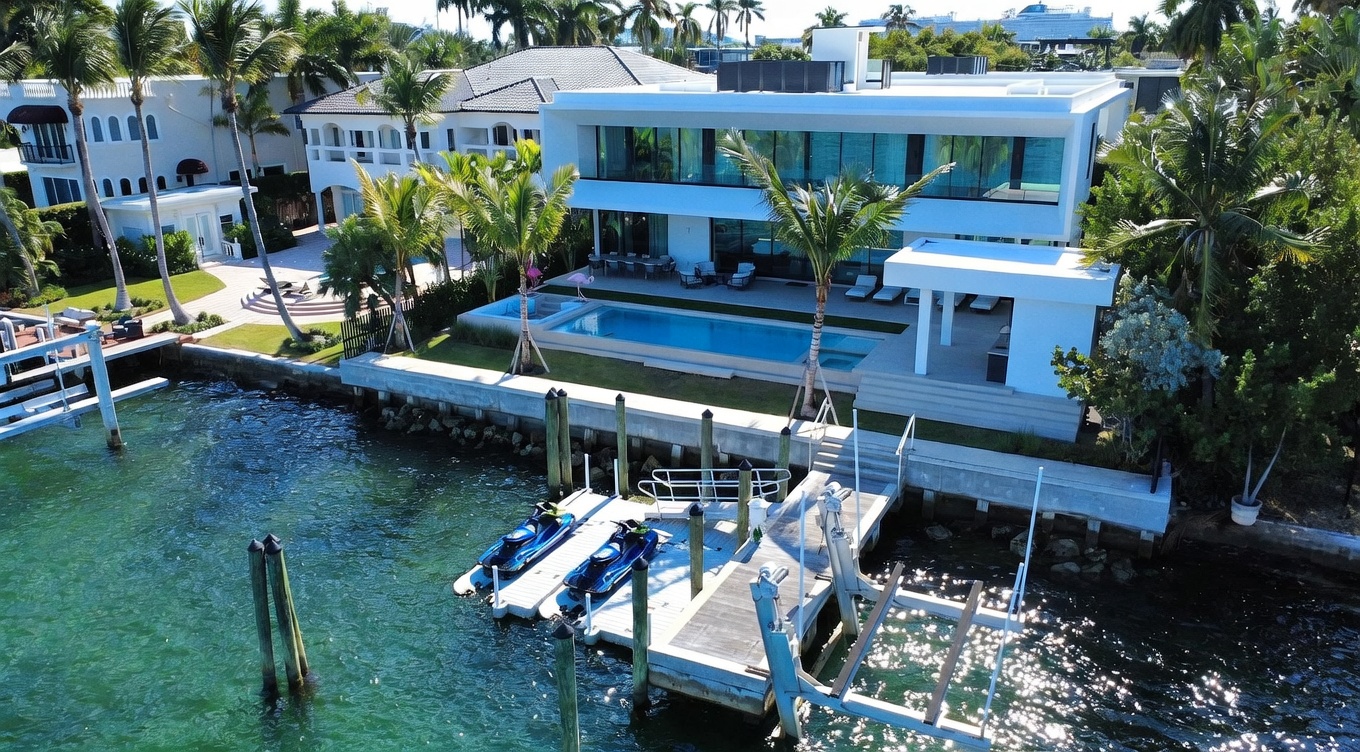 This high-angle aerial view captures a luxurious modern waterfront estate featuring a sleek, white multi-level residence with extensive glass walls. The property includes a private swimming pool, a manicured lawn, and a private dock equipped with two jet skis extending into the turquoise water. The scene conveys an upscale, resort-like atmosphere, highlighting the seamless integration between the contemporary architecture and the coastal environment.