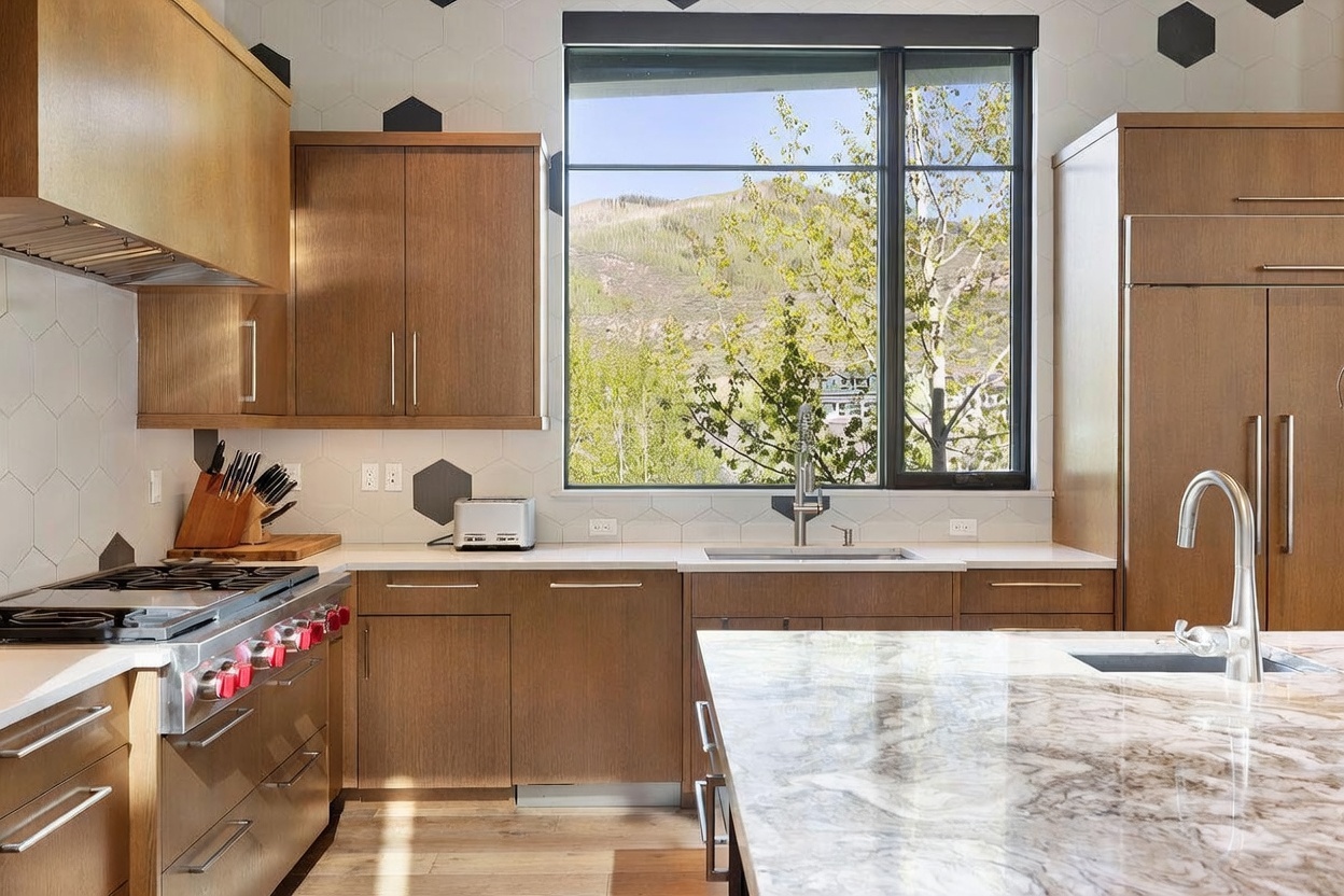 This modern kitchen features warm-toned wood cabinetry paired with light-colored stone countertops and a large central island. A prominent window above the sink provides a scenic view of the mountains and trees, while a professional-style gas range with a stainless steel hood serves as a focal point. The backsplash is adorned with a geometric hexagonal tile pattern, creating a sophisticated and contemporary aesthetic.