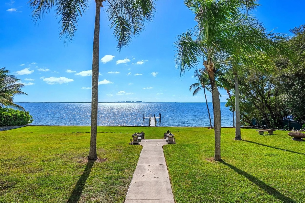 This expansive waterfront backyard features a lush green lawn leading directly to the water's edge, framed by tall palm trees. A central concrete pathway guides the eye toward a private dock extending into the blue bay, creating a serene and picturesque setting. The bright, sunny atmosphere highlights the open space and the unobstructed view of the horizon.