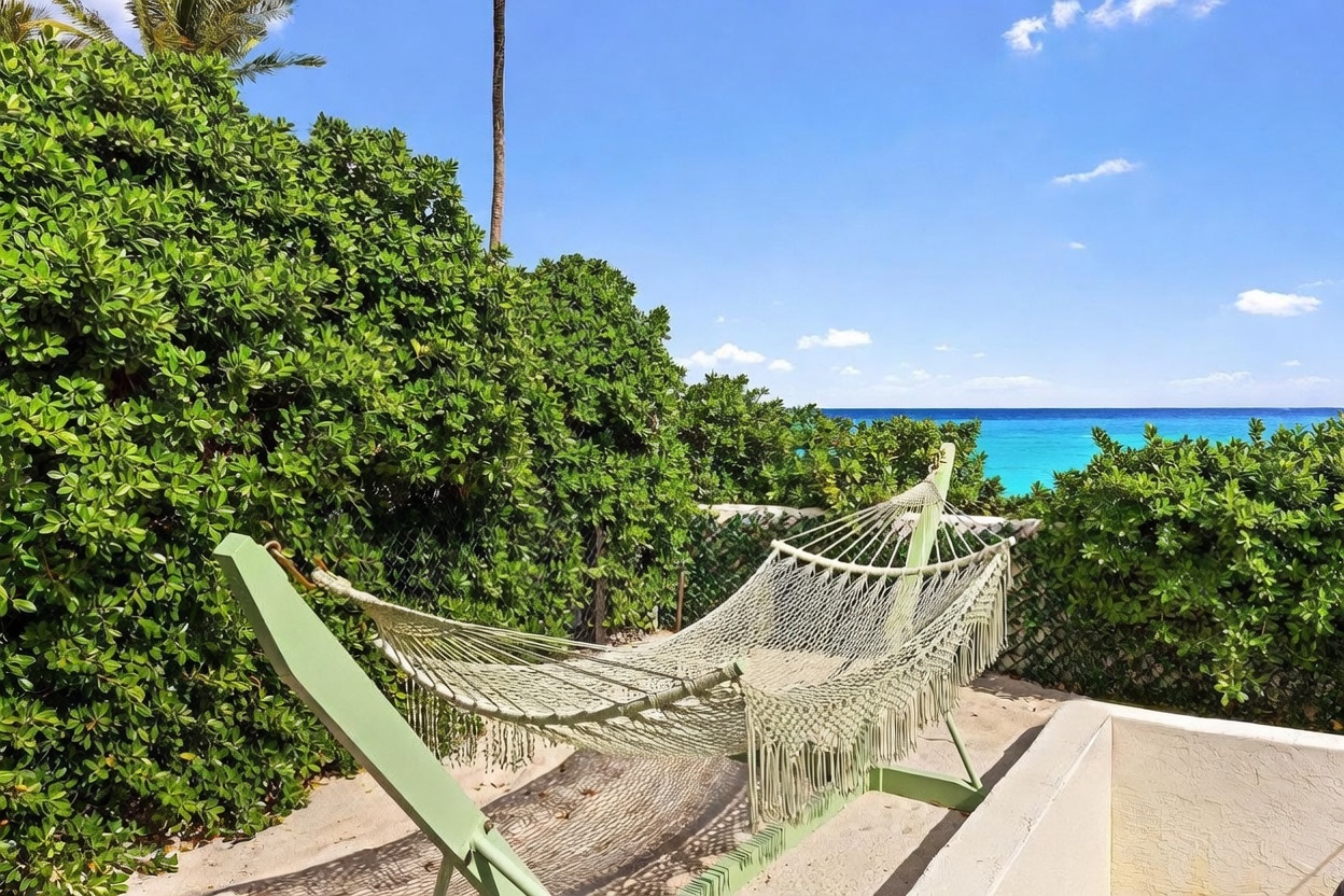 This serene outdoor space features a light green hammock set up on a paved patio, offering a relaxing spot to enjoy the coastal view. Lush green hedges provide privacy, while the backdrop showcases a vibrant turquoise ocean under a clear blue sky. The perspective is from a slightly elevated position, emphasizing the tranquil atmosphere and the direct connection to the seaside environment.
