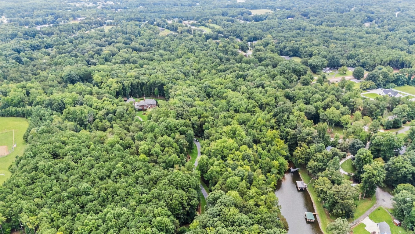 This high-angle aerial perspective captures a large, traditional brick estate nestled within a dense, lush forest. A winding paved driveway leads through the trees to the home, while a private body of water with a small dock sits to the right. The expansive property offers significant privacy and a secluded, natural setting.