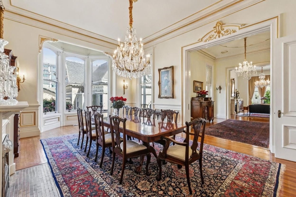 This elegant formal dining room features a large wooden table surrounded by ornate carved chairs, set beneath a grand crystal chandelier. The space is defined by classic architectural details, including crown molding, a bay window, and a wide doorway leading into an adjacent living area with a matching chandelier. A large, intricate patterned rug anchors the room, while a framed portrait and a wooden sideboard add a touch of traditional sophistication.