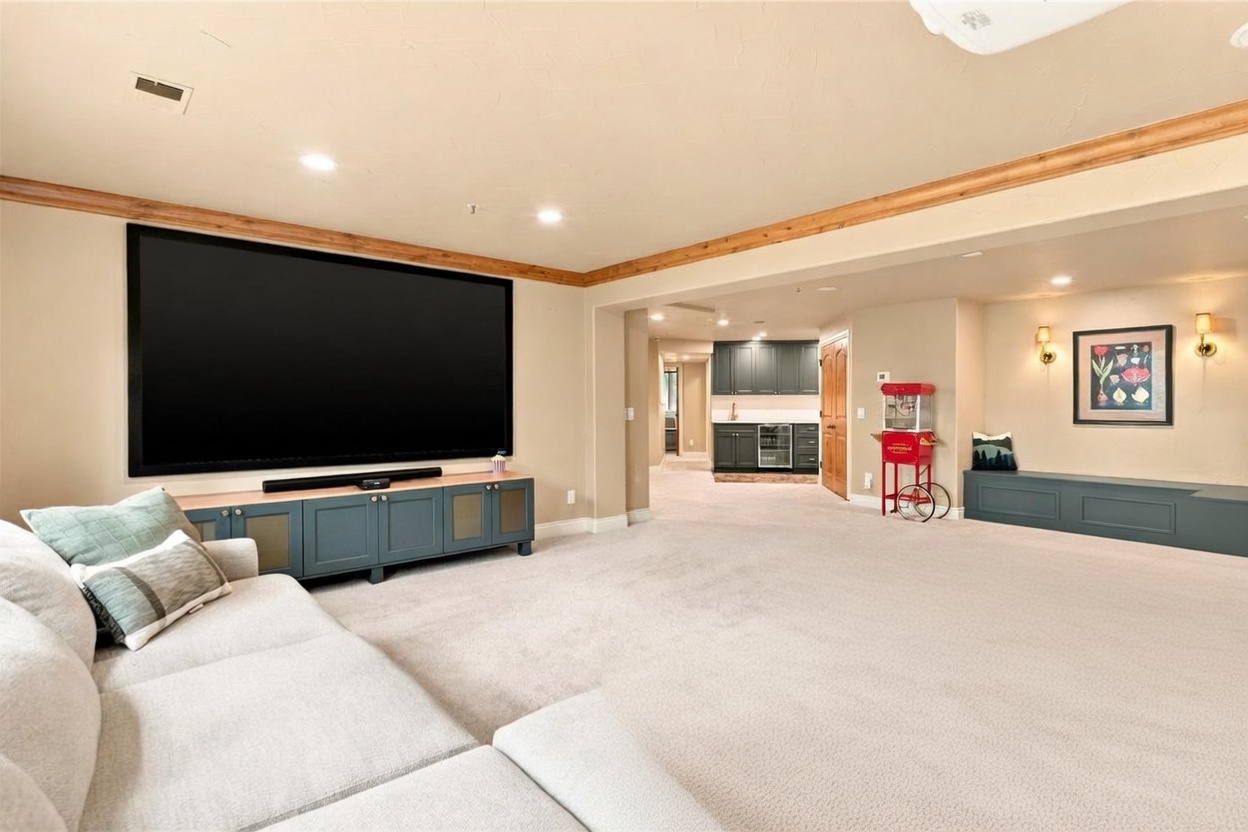 This spacious basement living area features a large wall-mounted television above a teal media console, creating a dedicated home theater setup. The room is furnished with a large, light-colored sectional sofa and includes a wet bar area in the background with dark cabinetry and a beverage refrigerator. A vintage-style popcorn machine adds a playful touch to the space, which is finished with neutral carpeting and warm recessed lighting.