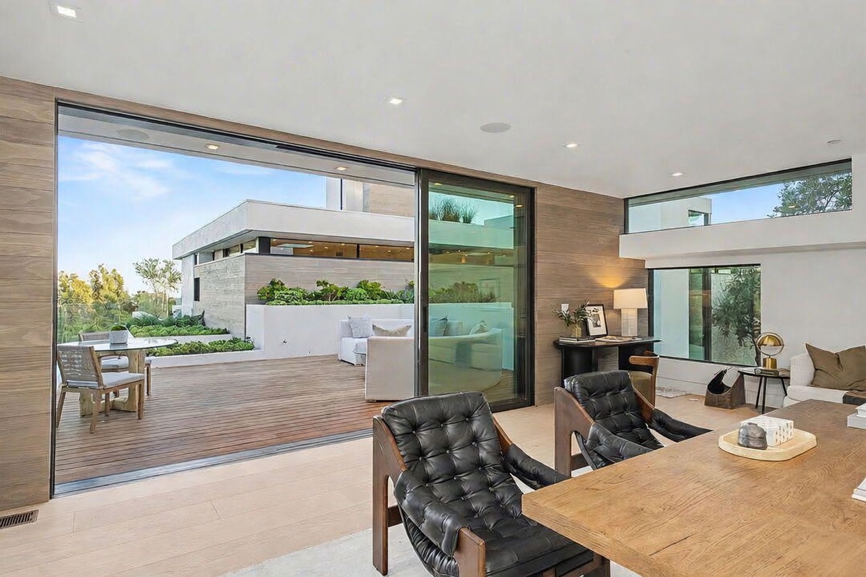 This contemporary living space features a seamless transition to an expansive outdoor deck through large sliding glass doors. The interior is characterized by warm wood-paneled walls, light-toned flooring, and a sophisticated dining area with black leather chairs. The open-concept design creates a bright, airy atmosphere that emphasizes indoor-outdoor living.