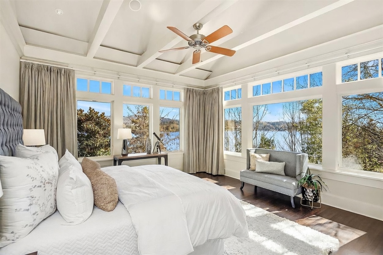 This luxurious primary bedroom features a coffered ceiling with a central fan and expansive windows that offer panoramic water views. The room is elegantly furnished with a plush bed, a sophisticated grey settee, and a console table, all set against dark hardwood floors and a soft white rug. The bright, airy atmosphere is enhanced by neutral tones and floor-to-ceiling curtains, creating a serene and high-end retreat.