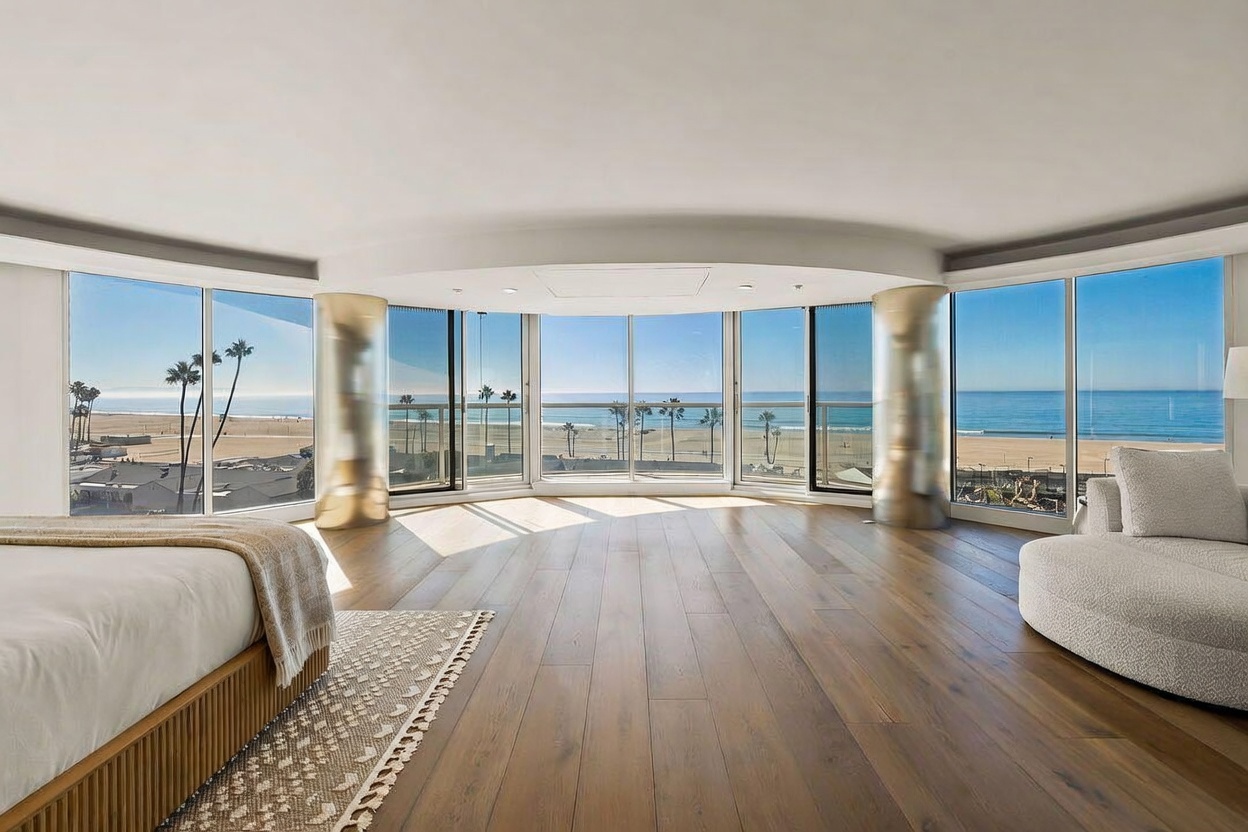This primary bedroom features a curved wall of floor-to-ceiling windows that provide an expansive, panoramic view of the beach and ocean. The room is anchored by light wood flooring and a neutral-toned bed, complemented by a curved sofa positioned to take advantage of the coastal scenery. Two metallic, cylindrical columns frame the central window section, adding a modern architectural element to the bright, airy space.