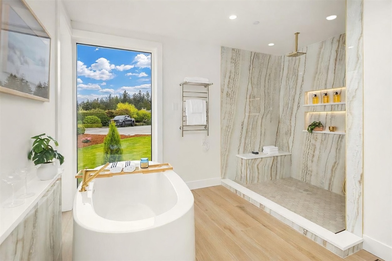 This luxurious primary bathroom features a freestanding soaking tub positioned next to a large window that provides a scenic view of the outdoors. To the right, a spacious walk-in shower is encased in glass and finished with striking, vertically-veined stone wall panels, integrated shelving, and a rainfall showerhead. The room is completed by light wood-toned flooring and a heated towel rack, creating a bright, spa-like atmosphere.