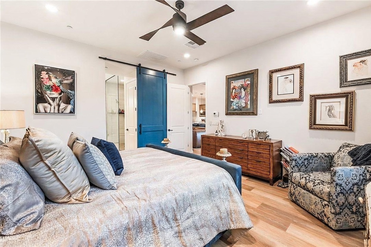 This primary bedroom features a large bed with a textured, metallic-toned comforter and several decorative pillows. The room is accented by a vibrant blue sliding barn door, a wooden dresser, and a patterned armchair, all set against light-colored walls and wood-look flooring. Multiple framed artworks adorn the walls, and a ceiling fan provides overhead lighting, creating a cozy and well-appointed living space.