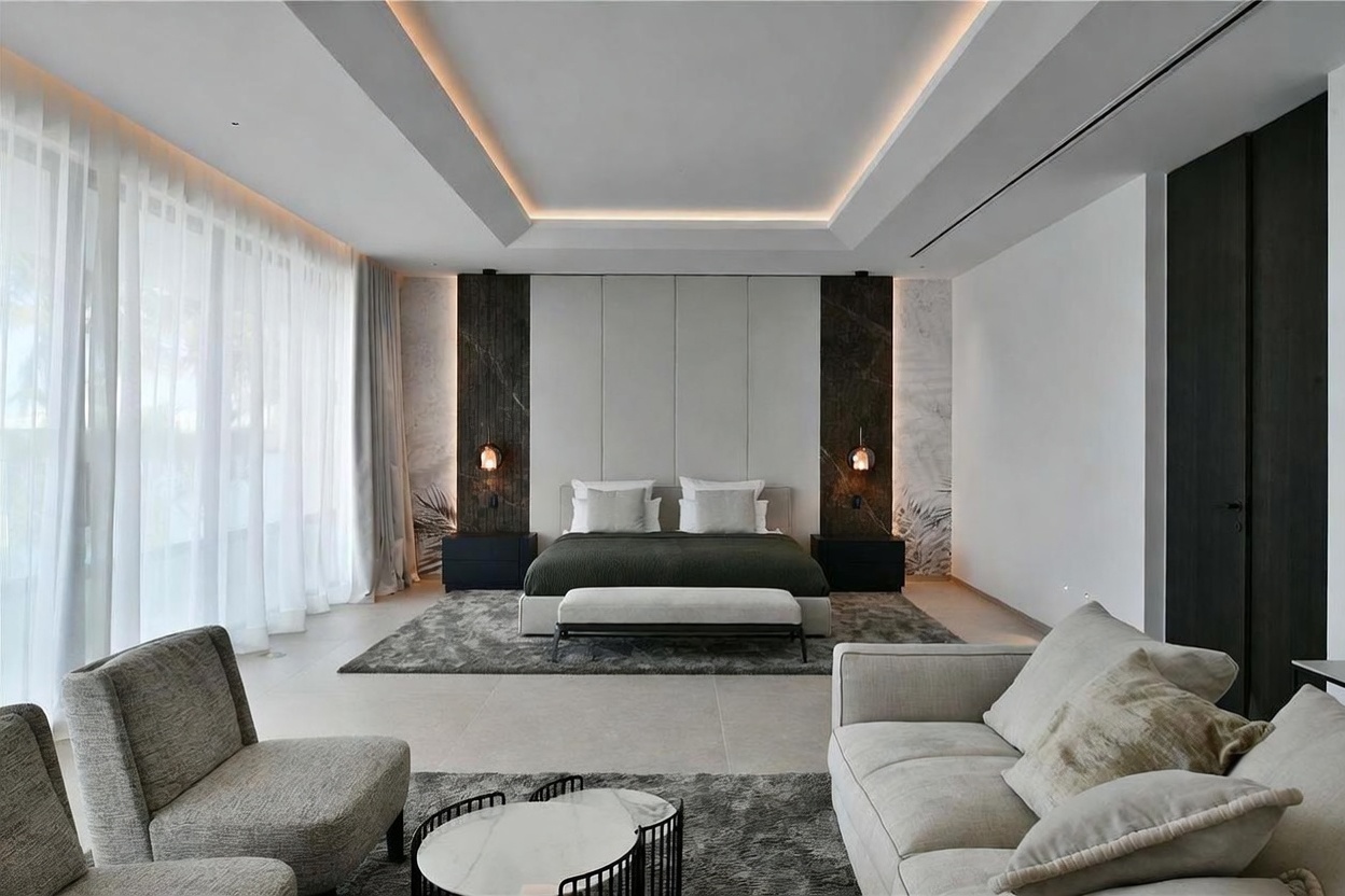 This expansive primary bedroom features a sophisticated, modern aesthetic with a neutral color palette and clean lines. The room is anchored by a central bed with a tall, upholstered headboard flanked by dark stone panels and integrated accent lighting, while a seating area with a sofa and armchairs occupies the foreground. Large windows draped in sheer curtains allow for abundant natural light, and the space is finished with a recessed tray ceiling and plush area rugs.