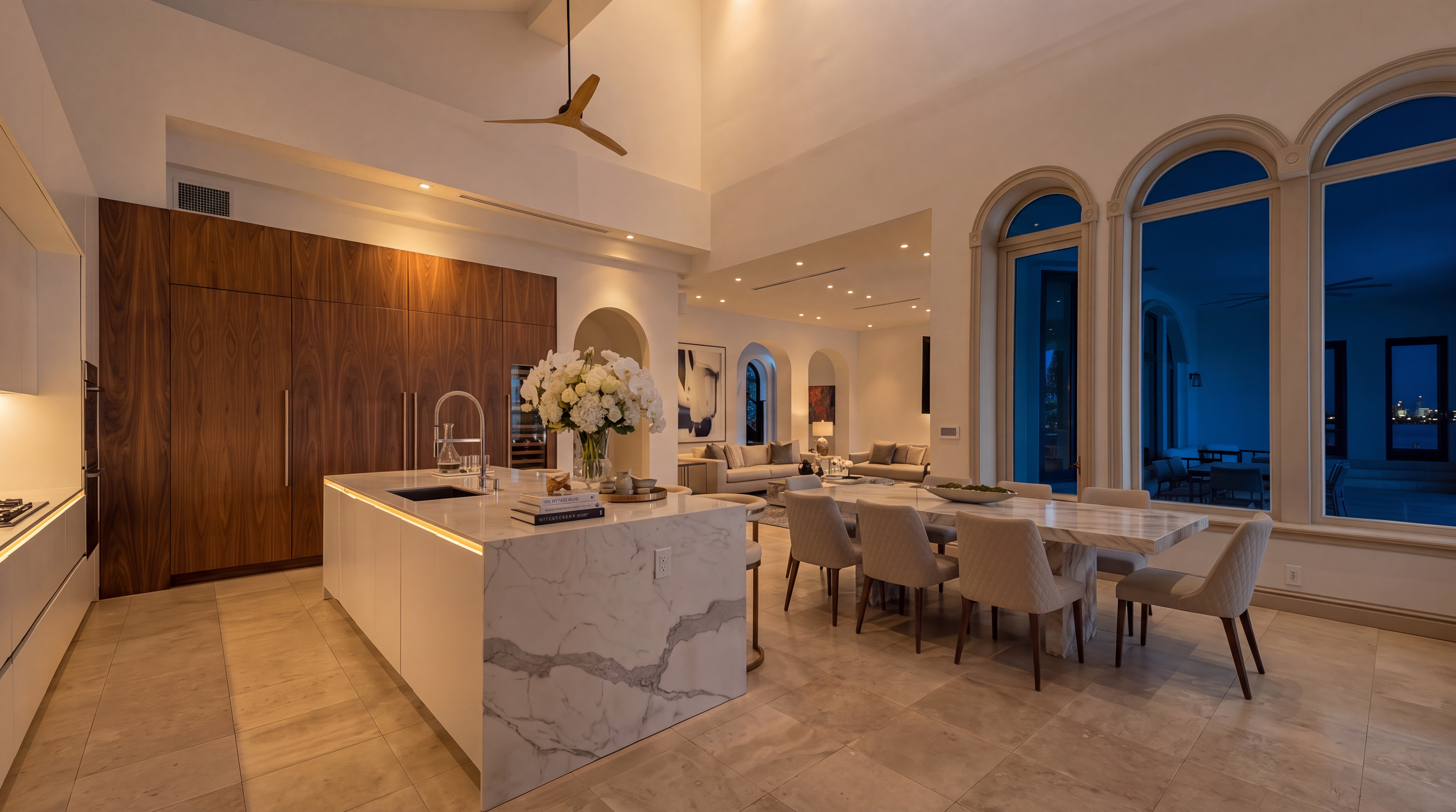 This stunning, expansive kitchen features a large marble-topped island and modern wood cabinetry, perfectly integrated into an open-concept layout that transitions into a formal dining area. Key highlights include high-vaulted ceilings with a statement fan, elegant arched floor-to-ceiling windows, and recessed lighting that creates an inviting, sophisticated atmosphere. Ideally designed for both cooking and entertaining, the space offers a seamless blend of luxury materials and contemporary functionality.