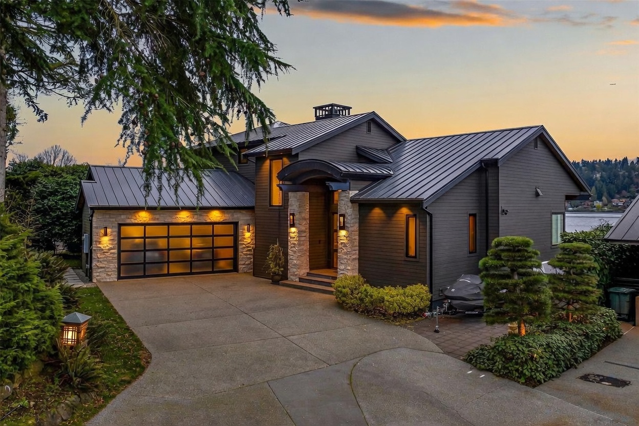 This modern home features a striking dark-toned exterior with a metal roof and stone accents around the garage and entryway. The front facade includes a multi-paneled glass garage door and a covered porch, all illuminated by warm exterior lighting at dusk. A concrete driveway leads up to the house, framed by manicured landscaping and a large tree in the foreground.