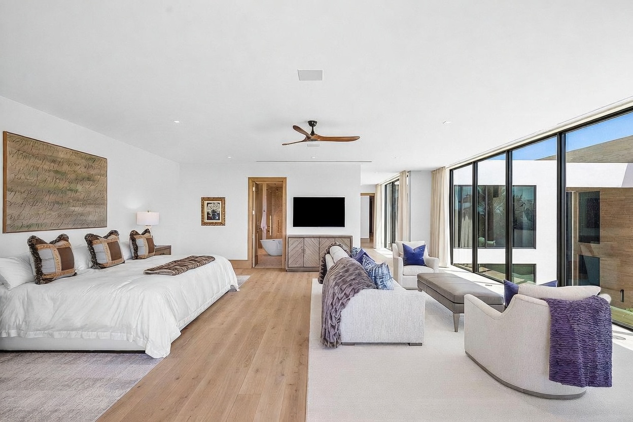 This expansive primary bedroom features a contemporary design with light wood flooring and a neutral color palette. The room includes a large bed with patterned accent pillows, a dedicated seating area with a sofa and armchairs, and floor-to-ceiling windows that provide abundant natural light and views of the exterior. A television is mounted on the wall above a wooden console, and a doorway leads to an adjacent bathroom, creating a seamless and luxurious living space.