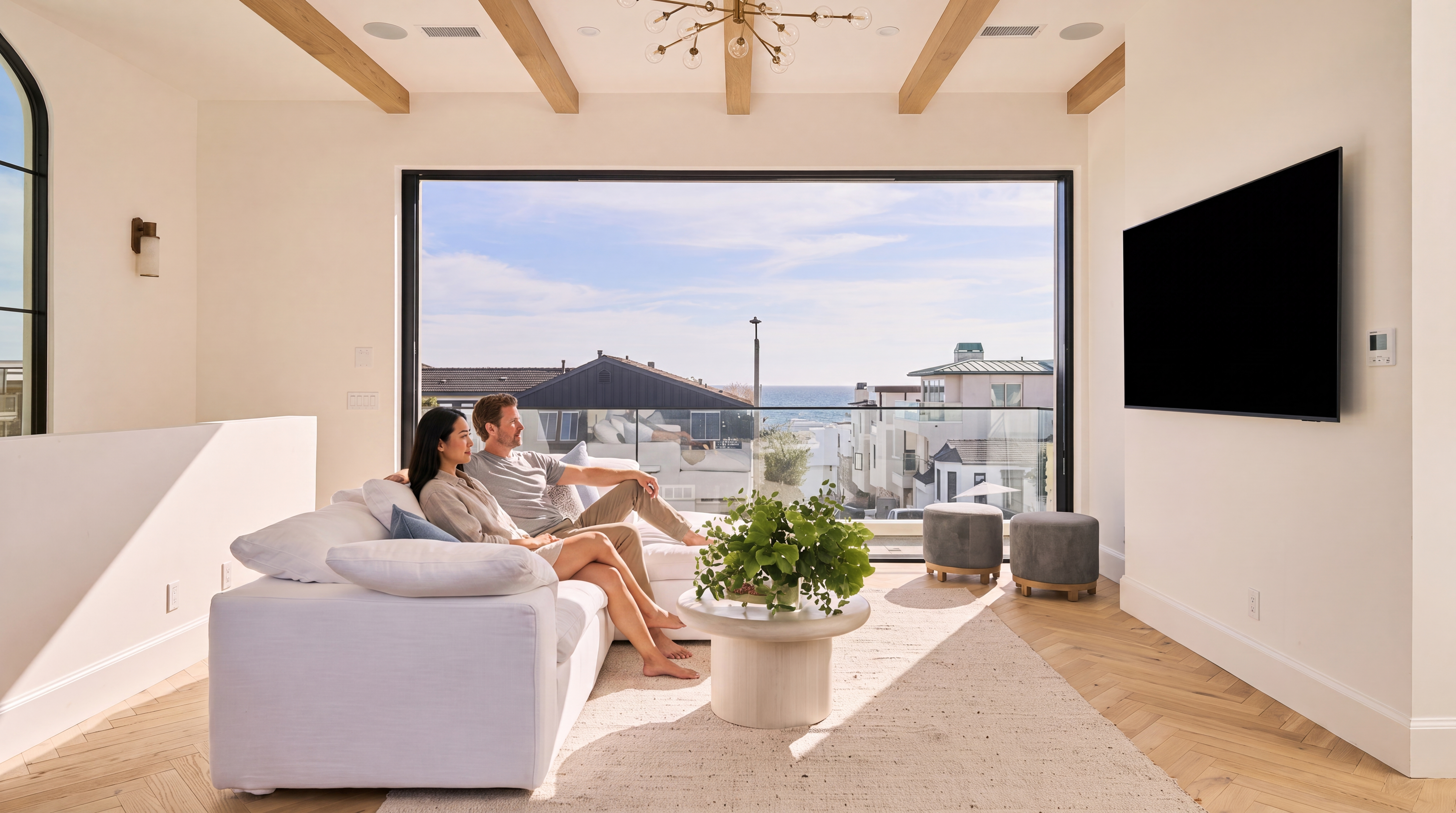 This living room features a bright, airy aesthetic with neutral tones, herringbone wood flooring, and exposed wooden ceiling beams. The space is anchored by a large white sectional sofa and a circular pedestal coffee table, with a prominent wall-mounted television, all set against a stunning floor-to-ceiling glass wall that offers expansive ocean views.