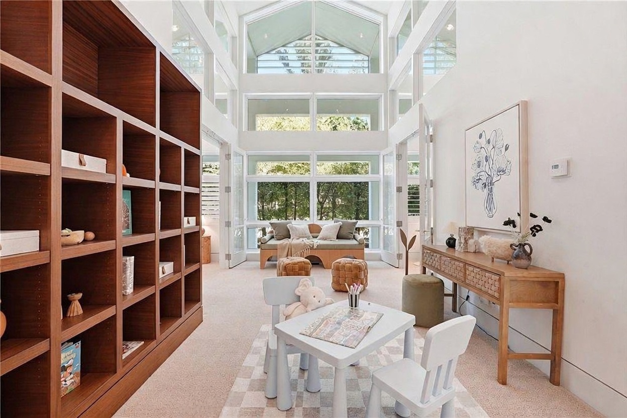 This bright and airy room serves as a versatile living or play space, characterized by its dramatic vaulted ceiling and expansive glass walls that flood the area with natural light. The room features a large, built-in wooden shelving unit on the left, a central children's activity table, and a cozy seating area with a daybed and ottomans near the rear windows. The neutral color palette and clean lines create a serene, inviting atmosphere suitable for a family-oriented home.