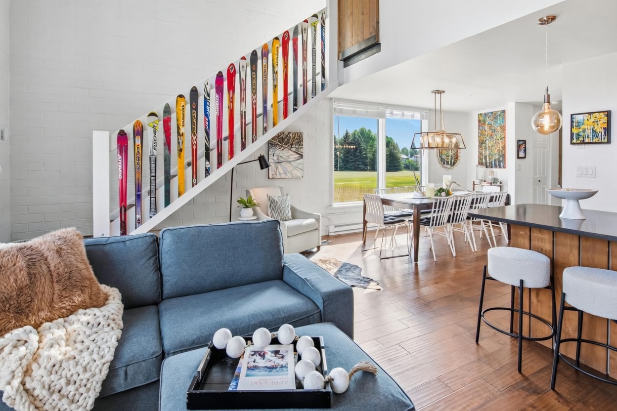 This open-concept living and dining area features a unique staircase railing decorated with a collection of colorful vintage skis. The space is anchored by a blue sofa and a matching ottoman, while the dining area beyond is illuminated by a modern chandelier and large windows overlooking a green lawn. The interior design blends rustic mountain charm with contemporary finishes, creating a bright and inviting atmosphere.