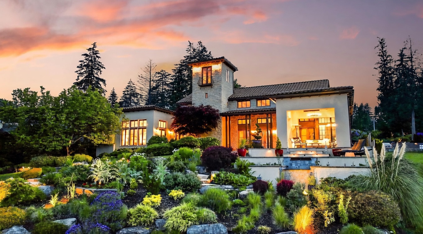 This stunning Mediterranean-style estate is captured at dusk, showcasing a multi-level design with a prominent stone tower and warm, inviting interior lighting. The foreground features a meticulously landscaped terraced garden filled with diverse shrubs, grasses, and flowers, leading up to a spacious covered patio area. The warm sunset sky provides a dramatic backdrop, highlighting the home's architectural character and its seamless integration with the lush outdoor environment.