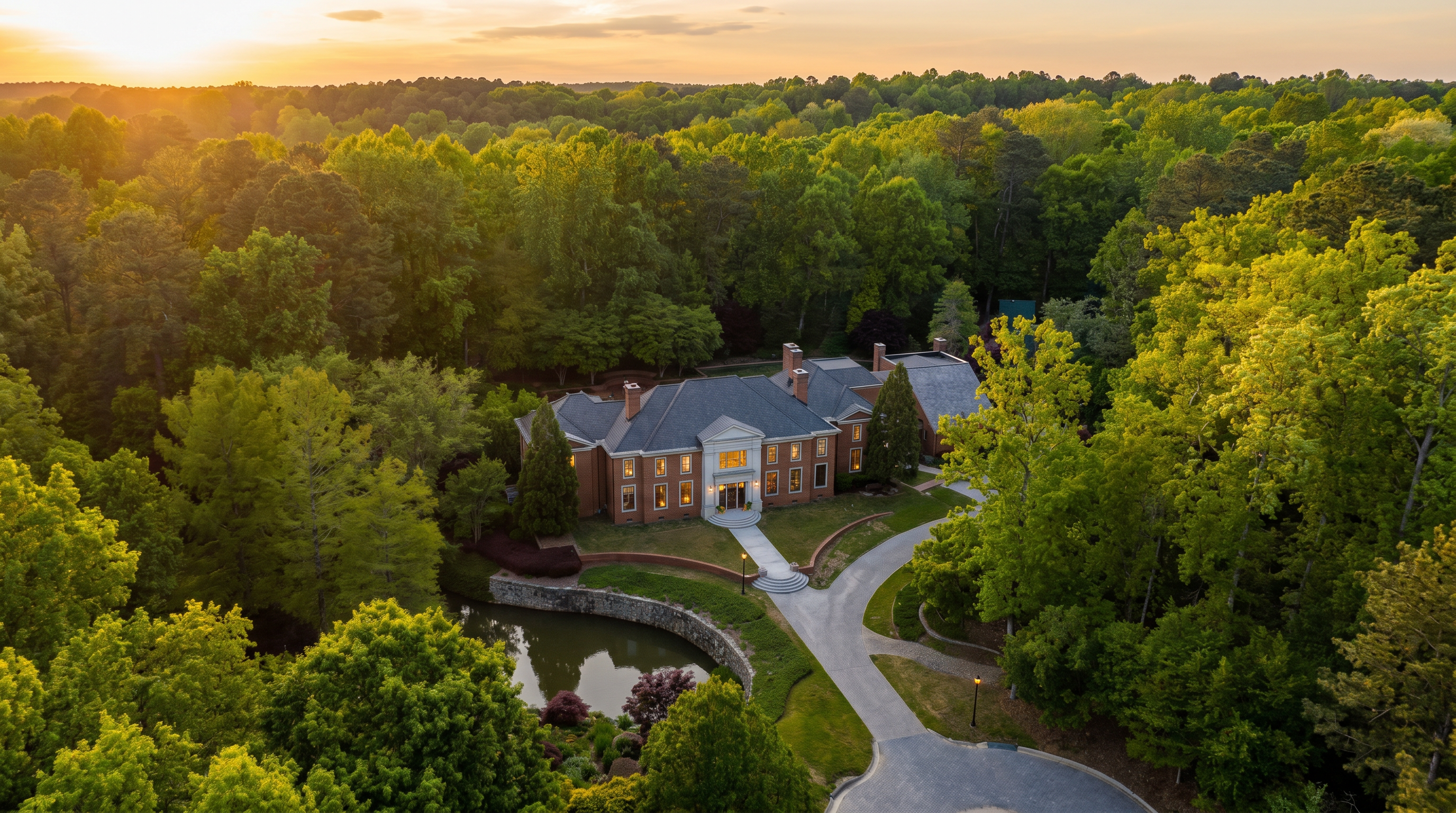 This high-angle aerial view captures a grand, multi-story brick estate nestled within a dense, lush forest. The property features a formal circular driveway, a manicured lawn, and a small, curved pond situated in the foreground. The architecture showcases a traditional aesthetic with a prominent central entrance and a multi-gabled roofline, emphasizing the home's secluded and expansive setting.
