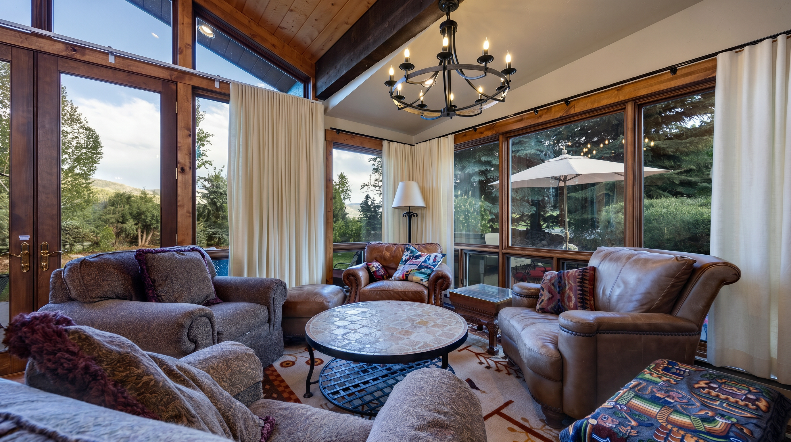 This cozy living room features a warm, rustic aesthetic with natural wood ceilings, a prominent chandelier, and comfortable, textured leather and upholstered seating arranged around a circular coffee table. Large windows surround the space, offering beautiful outdoor views and inviting an abundance of natural light to reach the interior. The room's integration of plush fabrics and wood accents creates a welcoming and inviting atmosphere typical of a upscale mountain retreat.