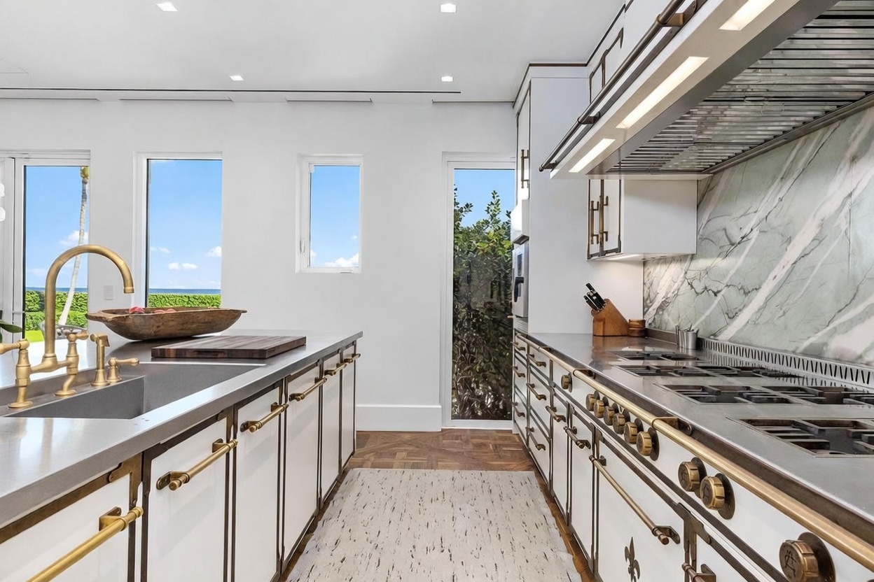 This high-end kitchen features white cabinetry accented with brass hardware and a striking green-veined stone backsplash. A large central island with a deep sink and a wooden cutting board sits opposite a professional-grade range, while tall windows provide natural light and views of the outdoors. The space is finished with patterned wood flooring and a neutral area rug, creating a sophisticated and functional culinary environment.