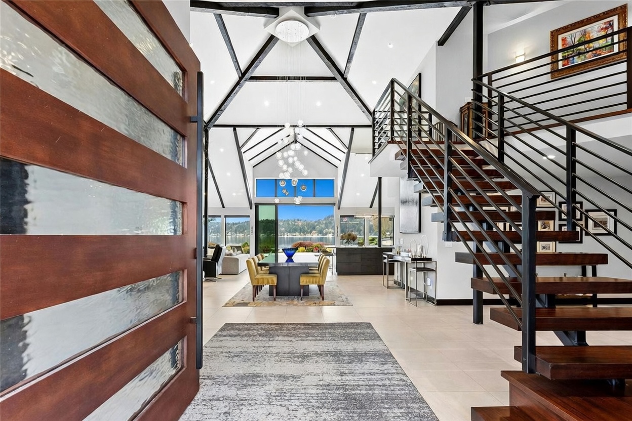 This striking entryway offers a dramatic perspective looking through a modern home toward a scenic lake view. The space features a soaring vaulted ceiling with exposed black beams, a grand floating staircase with metal railings, and a contemporary dining area that seamlessly transitions to the outdoors. The combination of warm wood tones, sleek industrial elements, and expansive glass creates a luxurious and airy atmosphere.