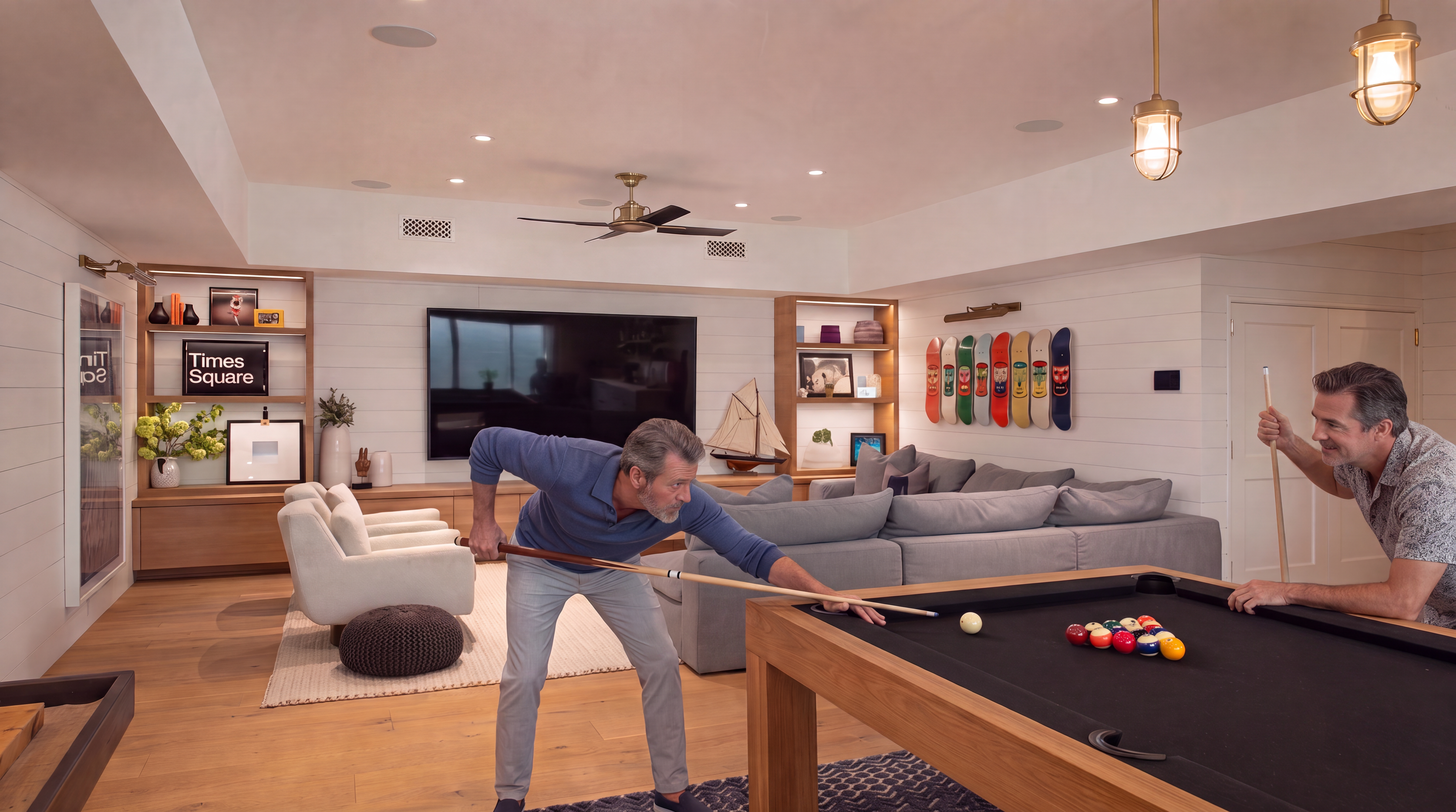 This spacious, open-concept living room features light wood flooring, white shiplap walls, and a large wall-mounted television centered above a long, low-profile wood media console. The room is furnished with a comfortable grey sectional sofa and two light-colored armchairs, complemented by a pool table in the foreground. Decorative elements include built-in shelving units, a collection of colorful skateboards mounted on the wall, and warm pendant lighting, creating a relaxed and inviting atmosphere.