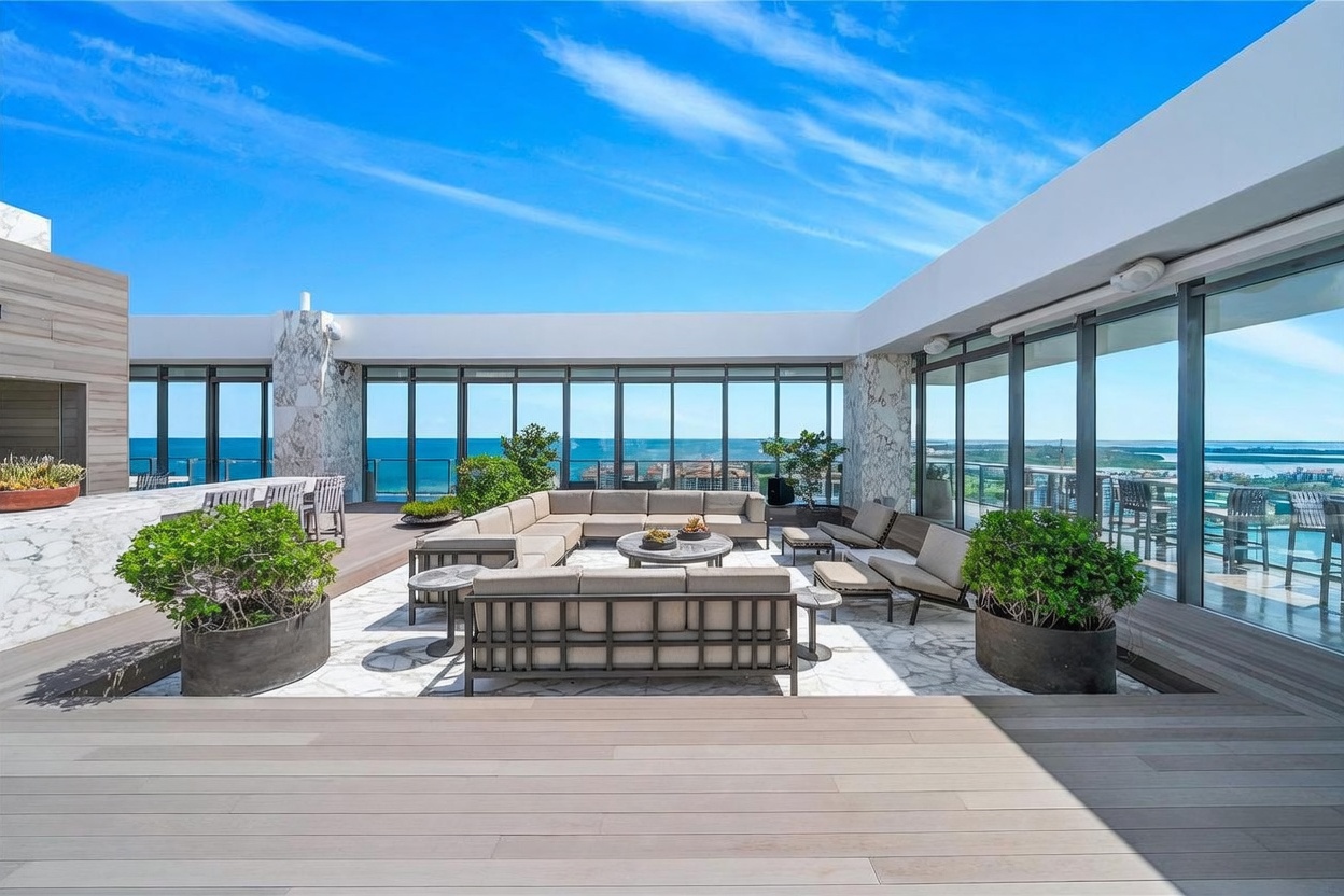 This expansive rooftop terrace features a sophisticated outdoor lounge area with modular seating arranged around a central circular table. The space is framed by floor-to-ceiling glass walls that offer panoramic ocean views, while large potted plants add a touch of greenery to the stone-tiled patio. A wide wooden deck in the foreground provides a warm contrast, creating an inviting and luxurious setting for outdoor entertaining.