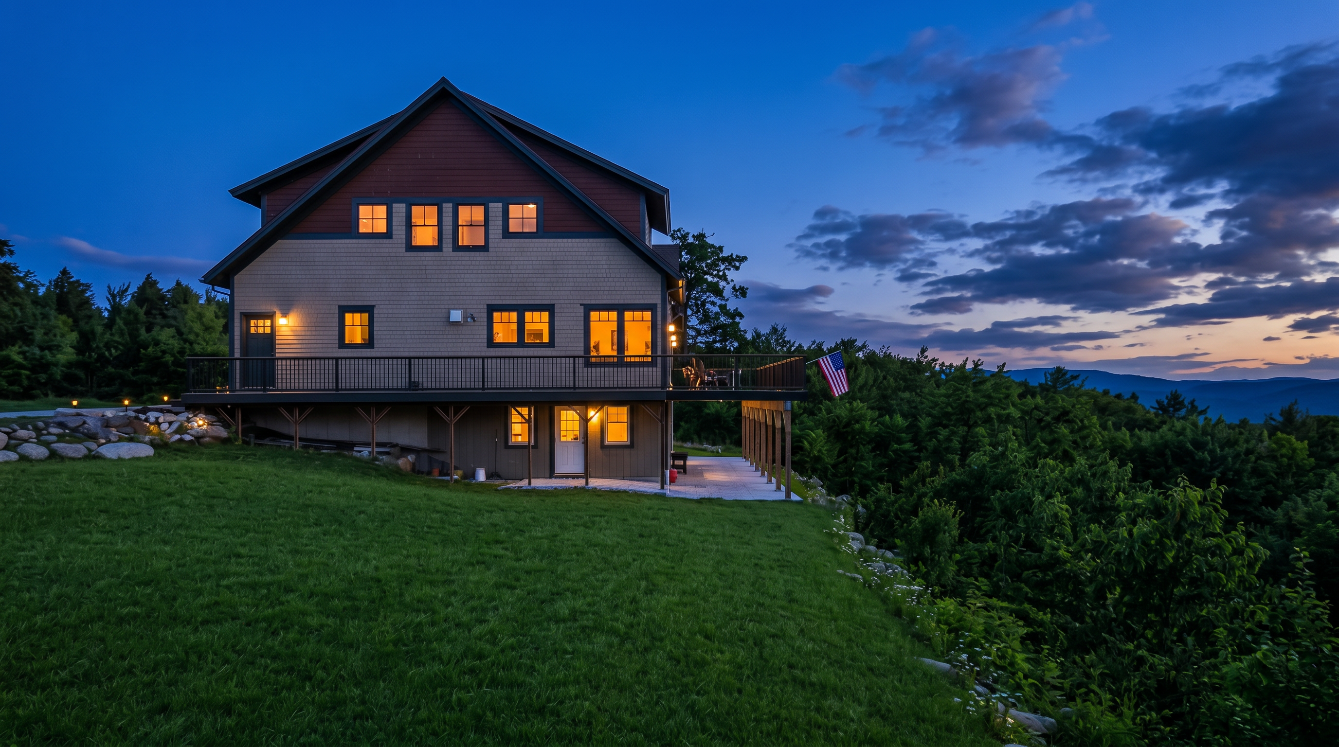 This exterior rear view showcases a multi-story home featuring a blend of brown shingle siding and matching wood-tone gables. The residence includes a full-length elevated deck with black railings and a ground-level covered patio area, all overlooking a vibrant, expansive green lawn. Its scenic position offers a peaceful environment while highlighting a well-maintained property with classic mountain-style architecture.