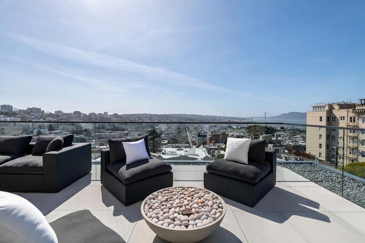 This expansive rooftop terrace offers a panoramic view of the city skyline and the Golden Gate Bridge in the distance. The space is furnished with comfortable dark-toned outdoor seating arranged around a circular stone-filled fire pit. A sleek glass railing provides an unobstructed vantage point, creating a sophisticated and airy atmosphere perfect for outdoor entertaining.