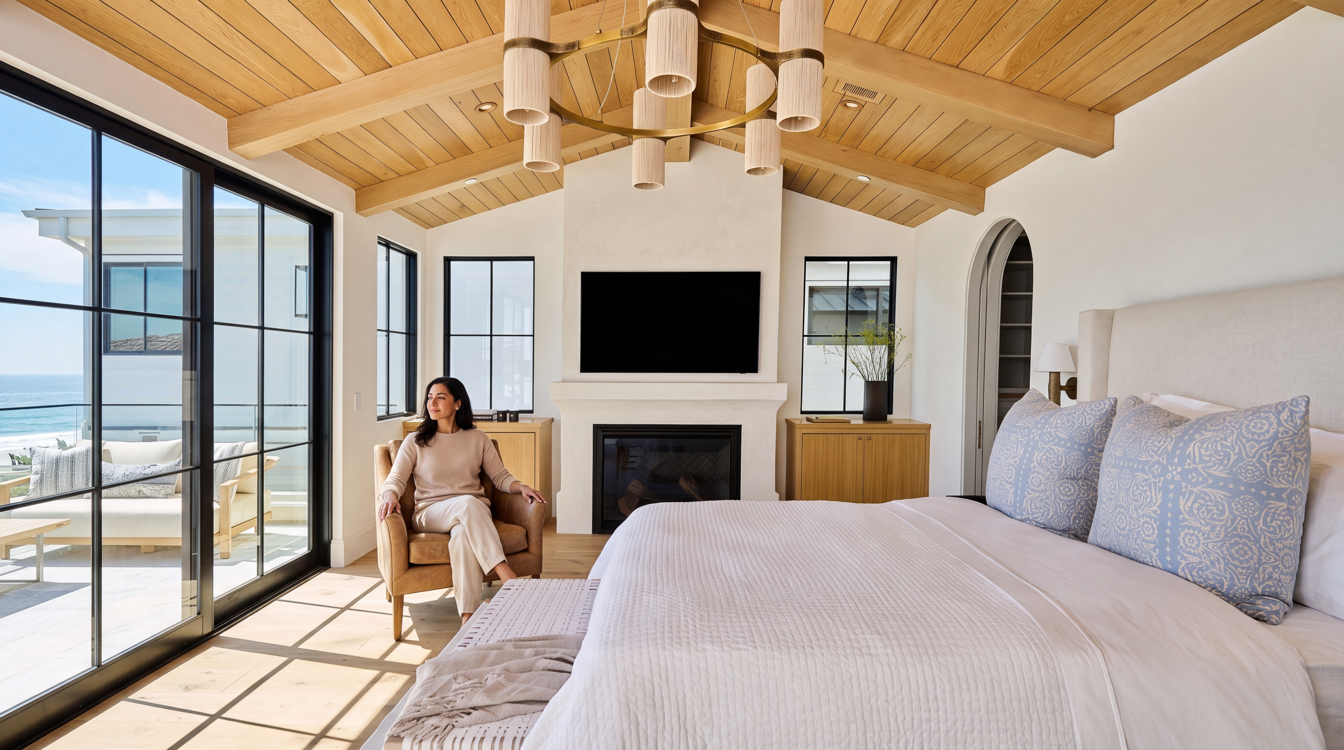 This luxurious primary bedroom features high vaulted wood-paneled ceilings, a cozy fireplace with an integrated flat-screen television, and expansive sliding glass doors that lead to a private balcony with ocean views. The room is decorated in a sophisticated, calm palette with light wood floors, a stylish chandelier, and a plush bed adorned with patterned pillows, creating an inviting and serene sanctuary.
