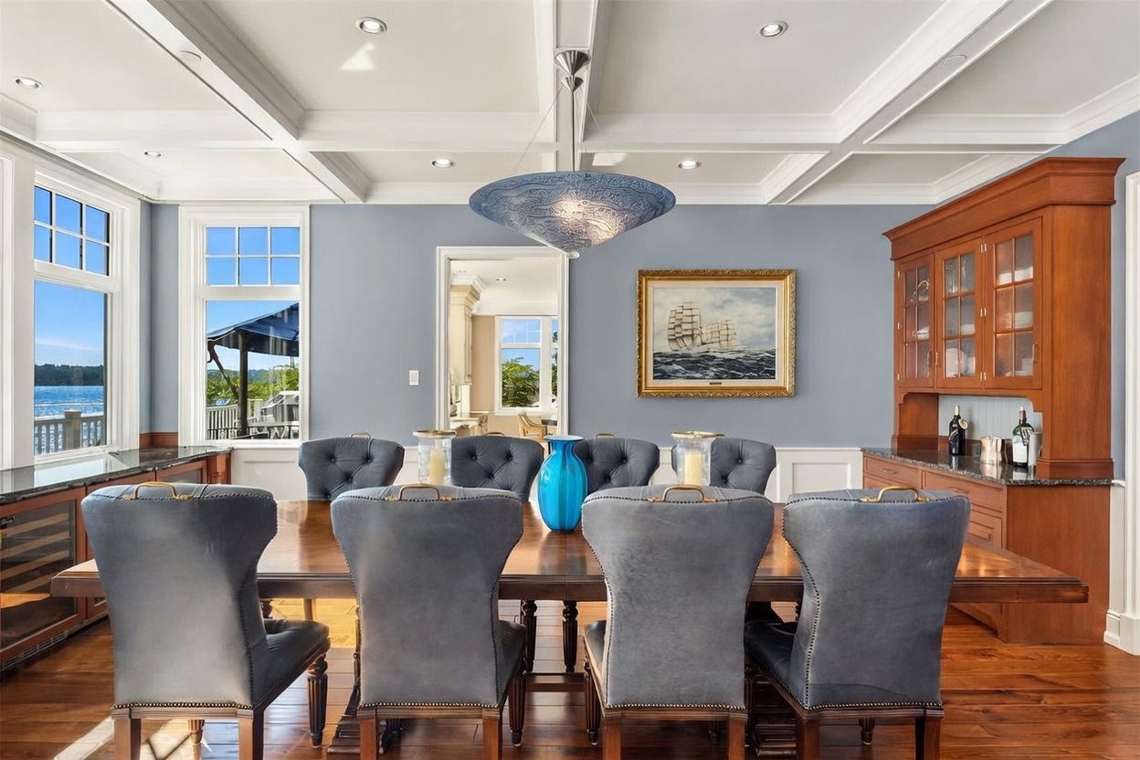 This elegant dining room features a large wooden table surrounded by eight plush, tufted grey chairs, set beneath a striking blue glass pendant light. The room is framed by a coffered ceiling, light blue walls, and a built-in wooden hutch, with large windows providing a scenic view of the water. The perspective is centered and symmetrical, capturing a sophisticated and inviting atmosphere perfect for formal gatherings.