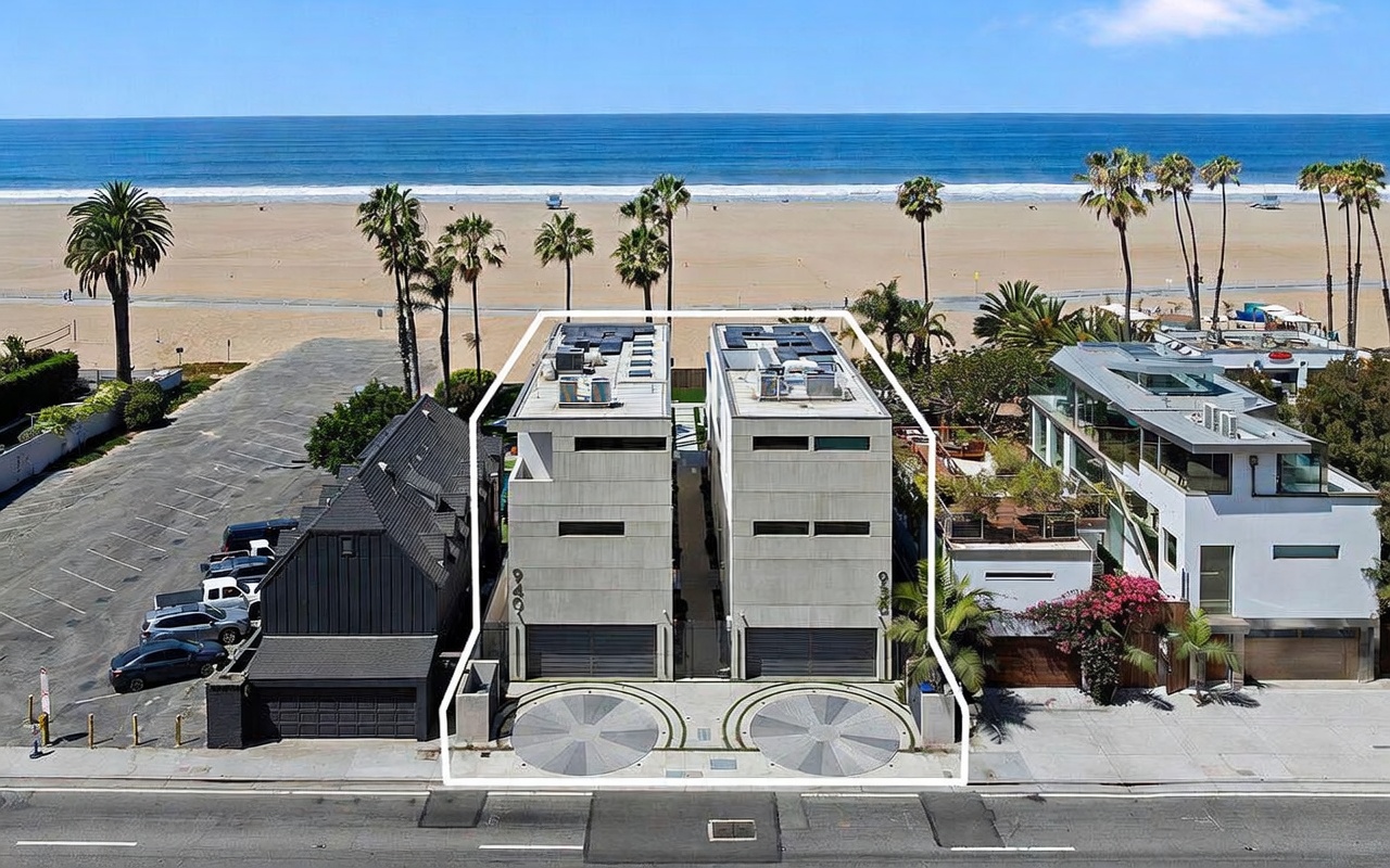 This aerial perspective captures two modern, multi-story residential structures situated directly along a sandy beach. The homes feature clean, geometric lines, flat roofs with solar panels, and integrated circular driveway turnarounds at the street level. The composition highlights the prime coastal location, showcasing the proximity to the ocean and the surrounding beach landscape.