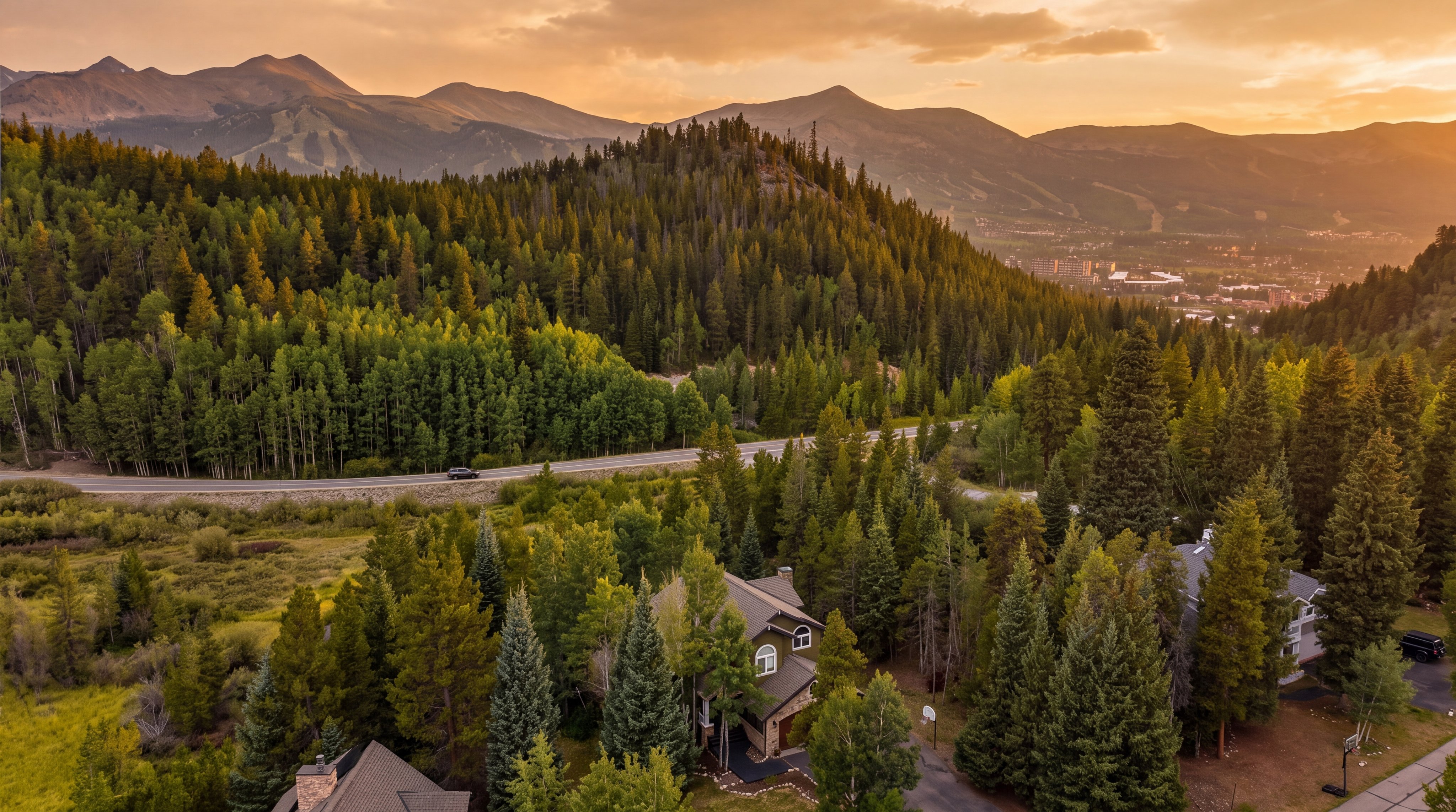 This breathtaking aerial photograph showcases a custom mountain home nestled within a dense, lush pine forest, offering an incredible sense of seclusion and natural beauty. The property features traditional architectural details with stone accents and a multi-pitched roofline that blends seamlessly into the stunning backdrop of dramatic, sun-drenched mountain peaks. With its elevated perspective, the home captures the serene essence of alpine living, perfectly complemented by the sweeping views of the surrounding valley and vibrant forest landscape.