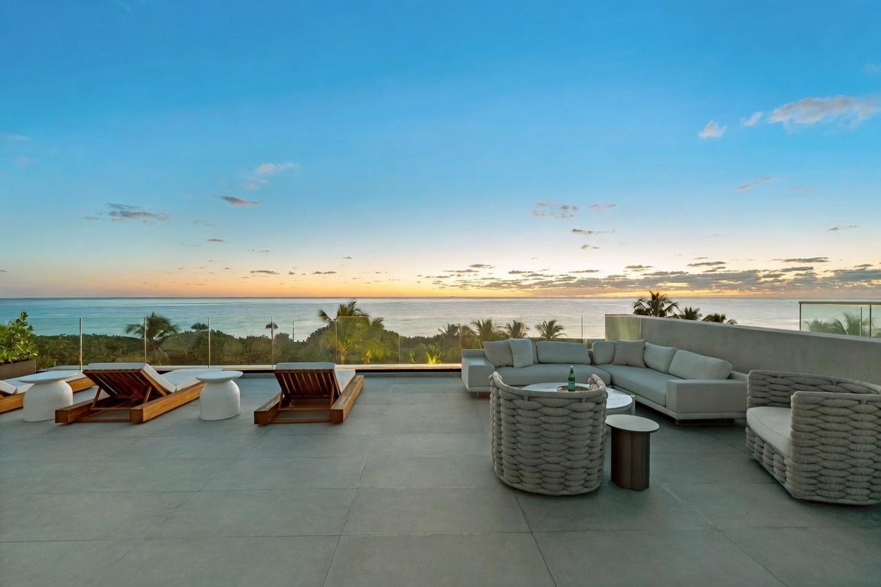 This expansive, modern terrace offers a luxurious outdoor living space with panoramic ocean views at sunset. The area is furnished with a large, light-colored sectional sofa, a woven circular chair, and several wooden lounge chairs arranged on a sleek, tiled deck. A glass railing provides an unobstructed view of the horizon, creating a seamless transition between the terrace and the coastal landscape.