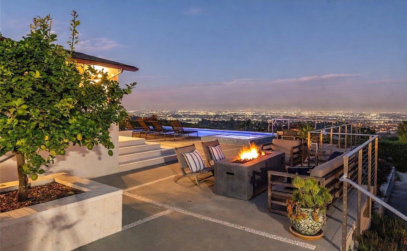 This expansive outdoor patio offers a luxurious setting for entertaining, featuring a modern fire pit as a central focal point. The space is furnished with comfortable lounge seating and overlooks an infinity-edge pool that blends seamlessly into a panoramic view of the city lights below. A mature lemon tree adds a touch of greenery to the foreground, while the overall atmosphere is one of sophisticated relaxation at dusk.
