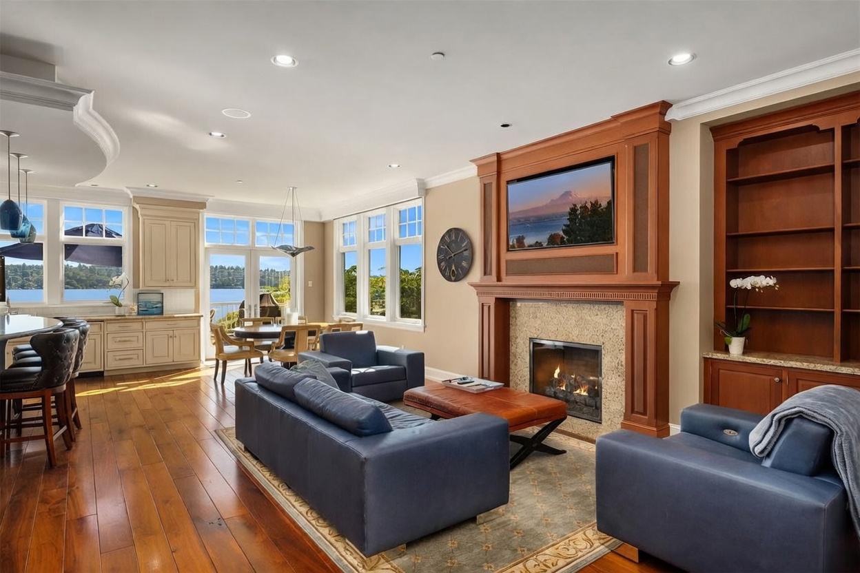 This spacious, open-concept living area features rich hardwood flooring, a grand fireplace with a built-in television, and custom shelving. The room flows seamlessly into a dining area and kitchen, all framed by large windows that offer a stunning, panoramic view of the lake. The overall aesthetic is sophisticated and inviting, perfectly blending comfort with high-end architectural details.