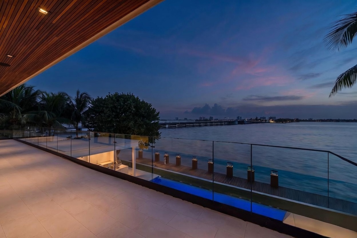 This expansive, modern balcony offers a panoramic view of the water at twilight, featuring a sleek glass railing that ensures unobstructed sightlines. The space is covered by a warm-toned wood-paneled ceiling with recessed lighting, creating a sophisticated atmosphere. Below, a private pool and a wooden dock with illuminated bollard lights extend toward the bay, highlighting the property's luxurious waterfront lifestyle.