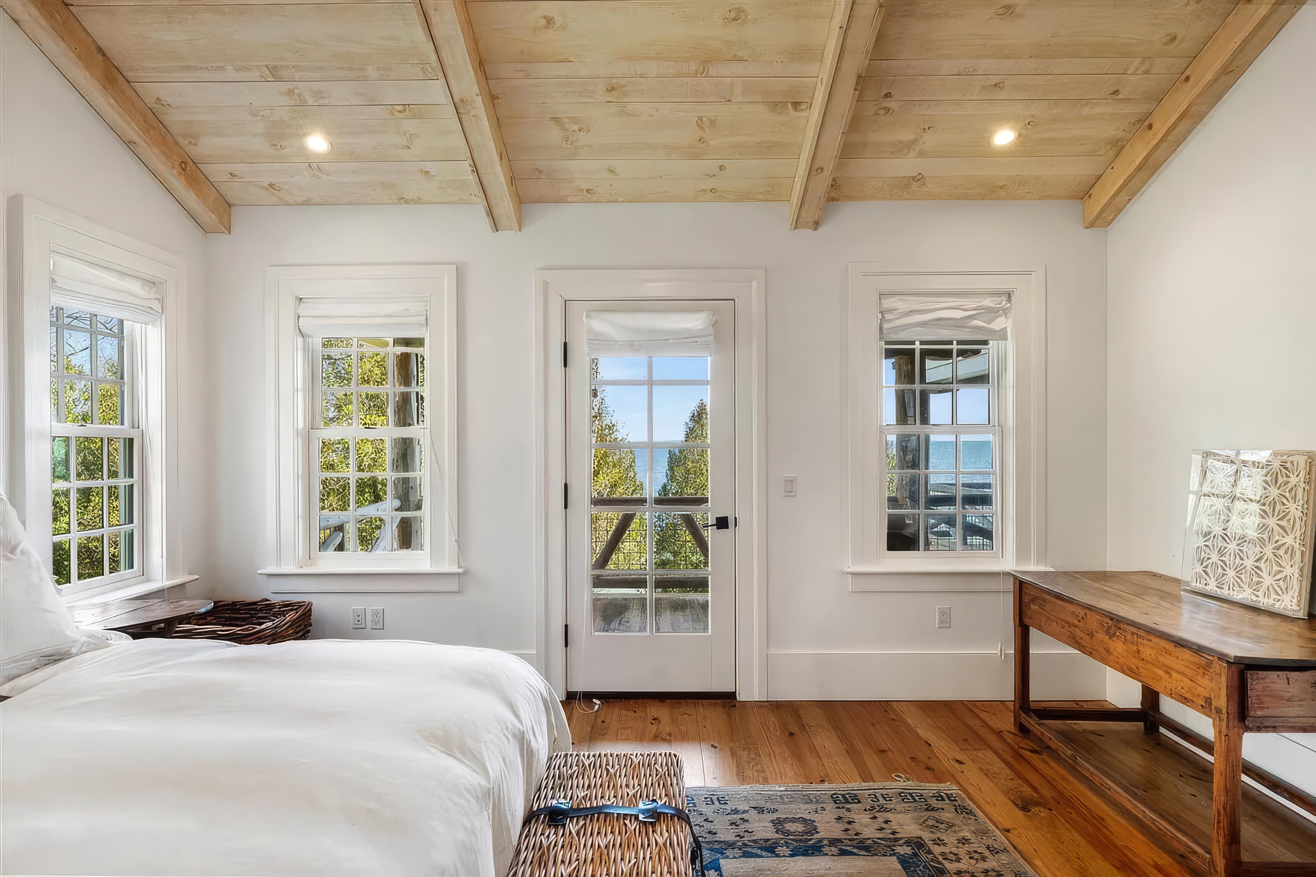 This bright and airy bedroom features a vaulted ceiling with exposed light wood beams and warm-toned wood flooring. A central glass-paned door flanked by two windows provides a scenic view of the outdoors and allows for abundant natural light. The room is furnished with a bed, a rustic wooden console table, and a woven bench, creating a serene and coastal-inspired atmosphere.