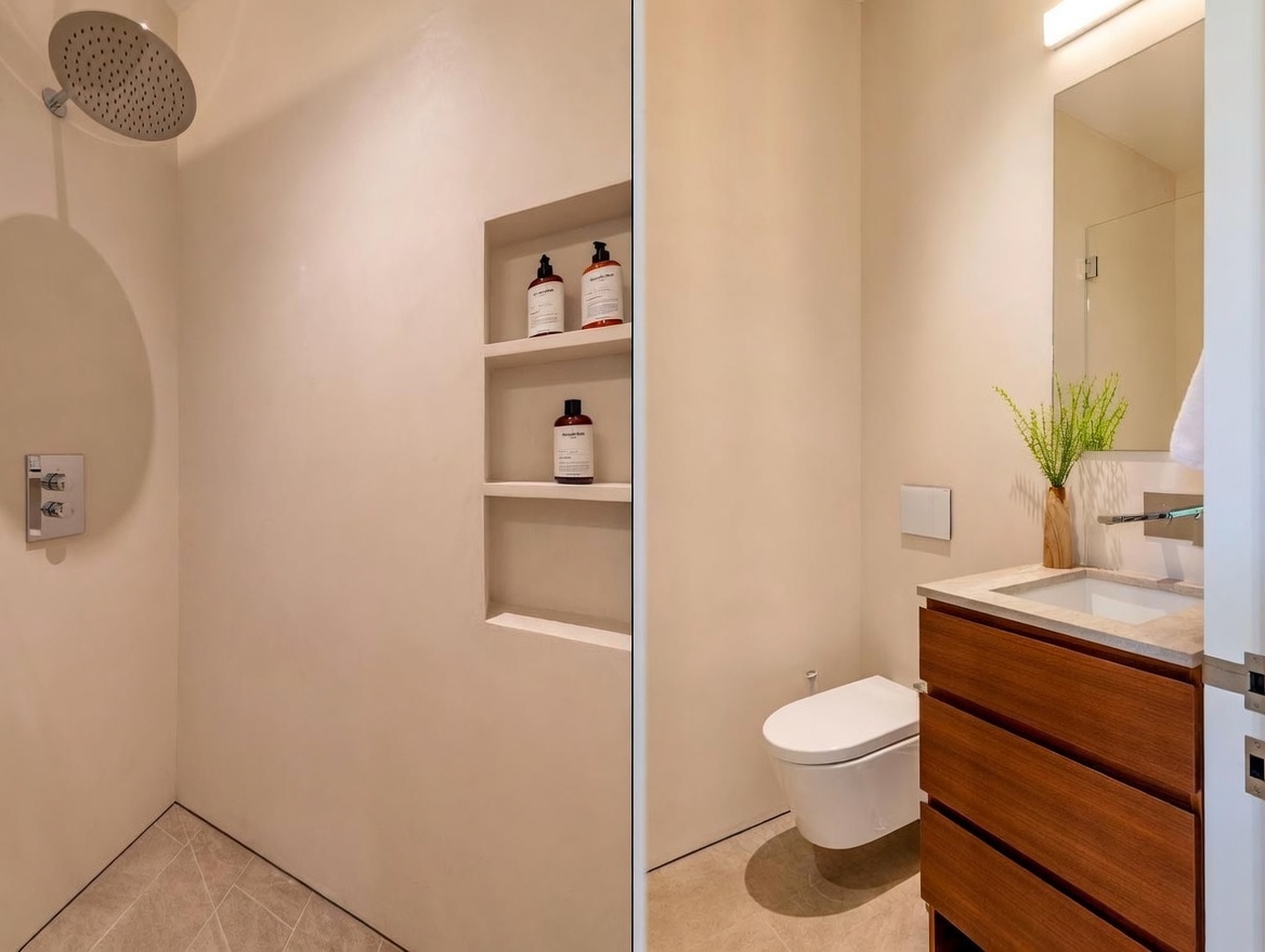 This split-view image showcases a modern, minimalist bathroom featuring a walk-in shower with a rainfall showerhead and recessed wall shelving on the left. The right side displays a wall-mounted toilet, a floating wood-toned vanity with a stone countertop, and a sleek mirror above. The space is characterized by neutral tones, clean lines, and a contemporary aesthetic.