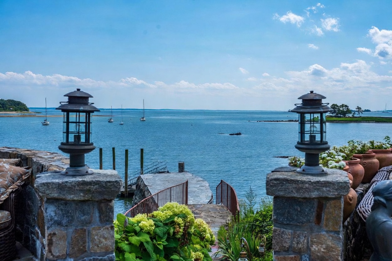 This elevated stone terrace offers a breathtaking, unobstructed view of a calm bay dotted with sailboats. The foreground features two classic lantern-style light fixtures mounted on stone pillars, framing a stone pathway that leads down to a private stone pier extending into the water. Lush green hydrangea bushes and terracotta planters add a touch of natural elegance to this serene, waterfront outdoor living space.